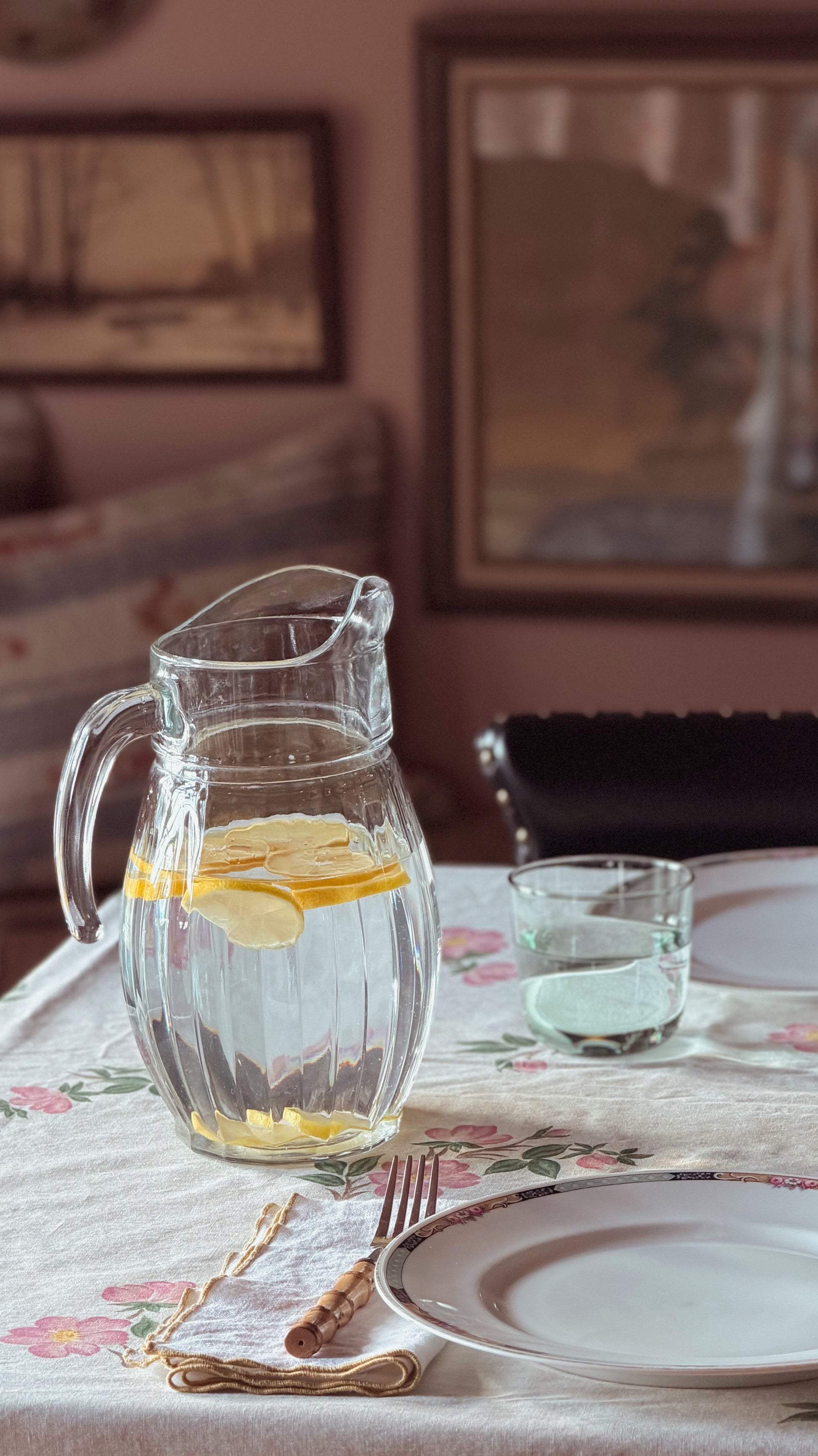 A clear glass pitcher filled with water and lemon slices sits on a floral tablecloth beside a matching glass, with plates and cutlery in a softly lit dining setting.