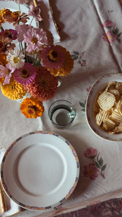 A styled tabletop scene with a floral tablecloth, a bouquet of pink, orange, yellow, and white flowers, an empty white plate with a floral rim, a small glass of water, and a Vintage Visnaud Limoges The Meuse Floral Oval Vegetable Bowl full of potato chips.