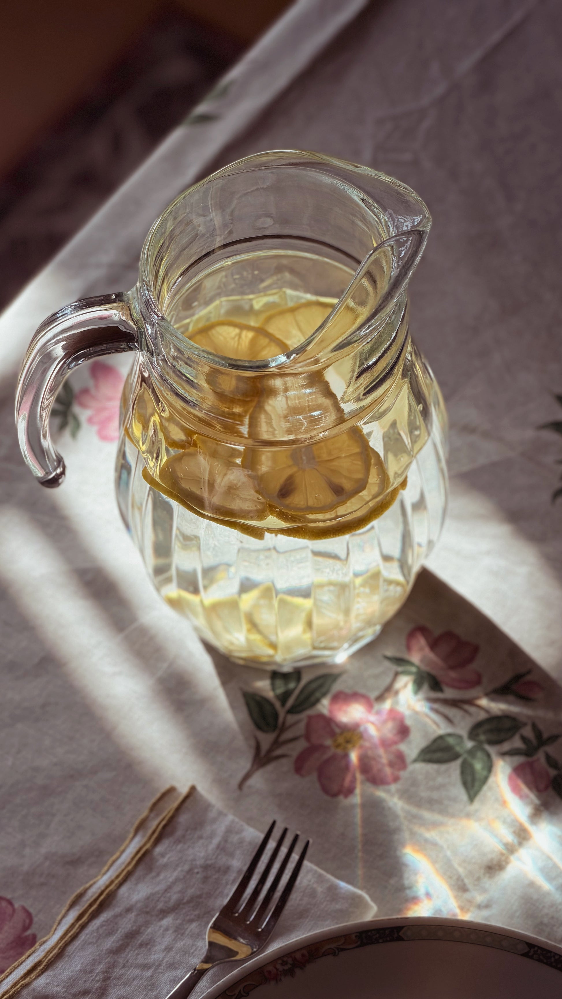 A clear glass pitcher filled with lemon slices and a light yellow beverage, placed on a floral tablecloth with warm sunlight and shadows.