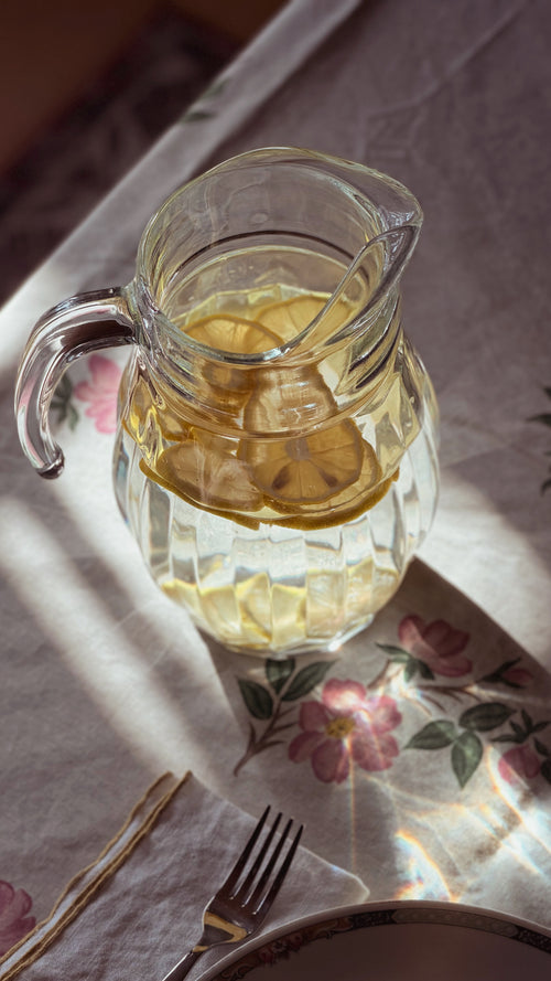 A clear glass pitcher filled with lemon slices and a light yellow beverage, placed on a floral tablecloth with warm sunlight and shadows.