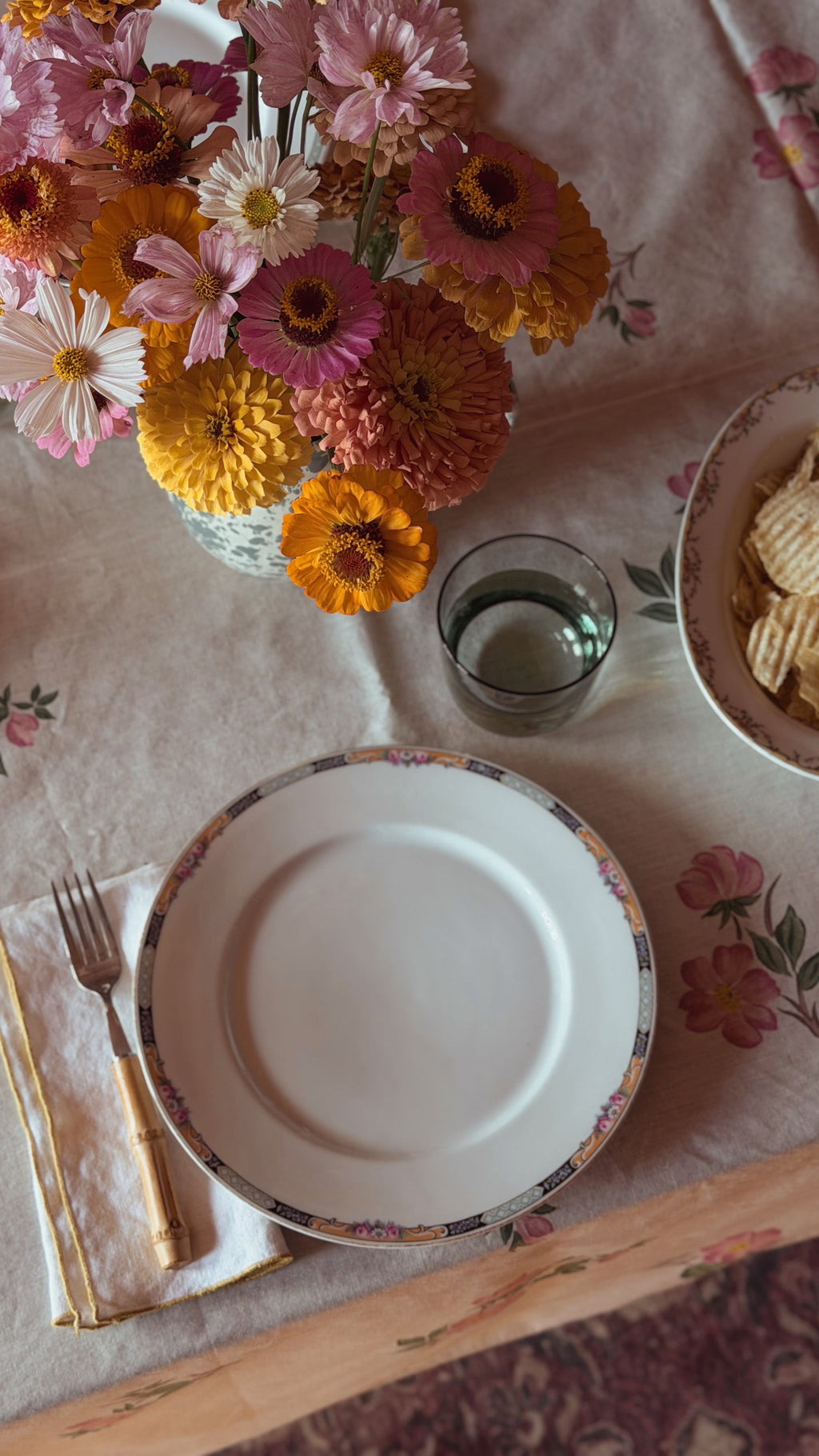 A styled tabletop setting featuring a Vintage KPM Germany Floral Porcelain Dinner Plate with a delicate floral border, accompanied by a gold-handled fork, a glass of water, and a bowl of chips on a floral tablecloth, with a vase of colorful daisies in the background.
