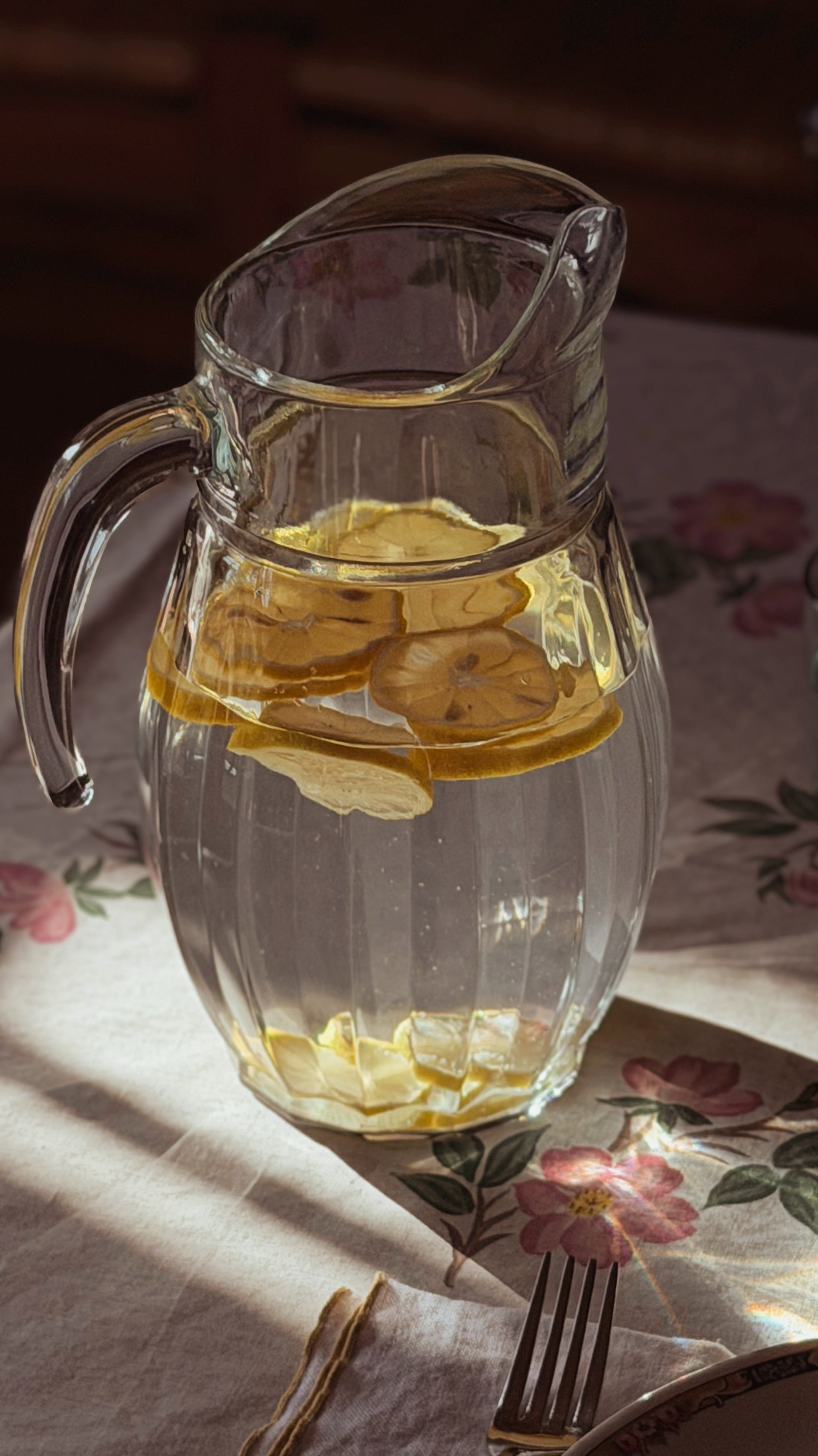 A Vintage Faceted Clear Glass Water Pitcher filled with lemon slices and water, set on a floral tablecloth with warm sunlight and shadows.