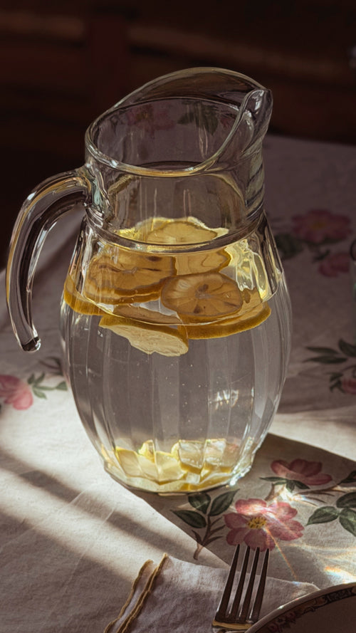 A Vintage Faceted Clear Glass Water Pitcher filled with lemon slices and water, set on a floral tablecloth with warm sunlight and shadows.