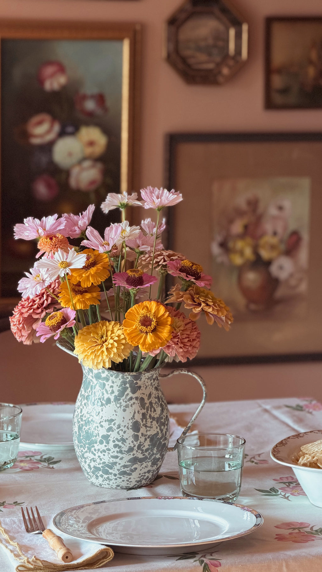 A rustic enamelware pitcher filled with a colorful bouquet of pink, yellow, and orange flowers, set on a floral tablecloth in a warmly lit dining room.