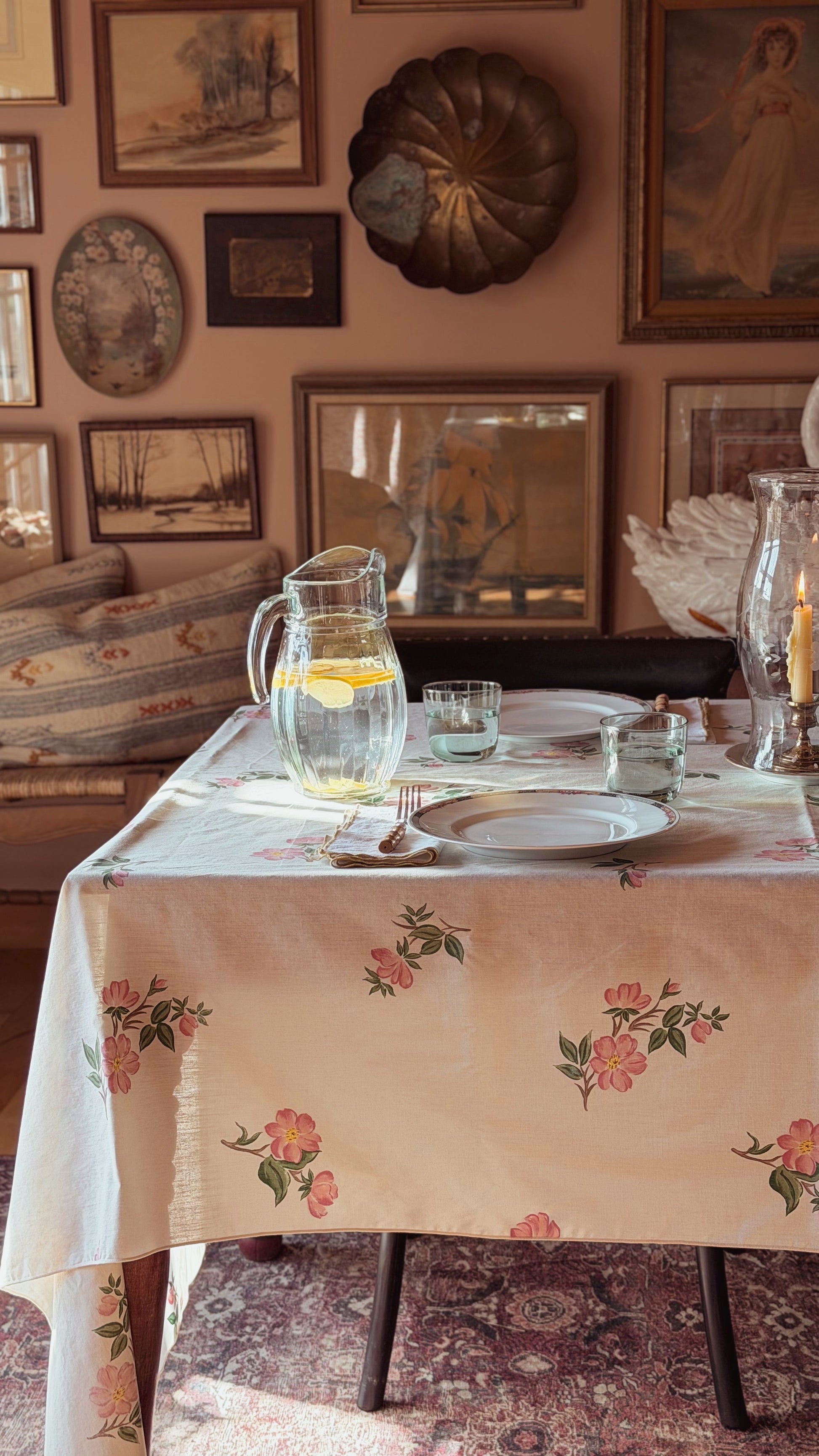 A floral tablecloth with pink and green rose patterns draped over a dining table, styled with a glass pitcher, water glasses, and plates in a warm, vintage-inspired room setting.