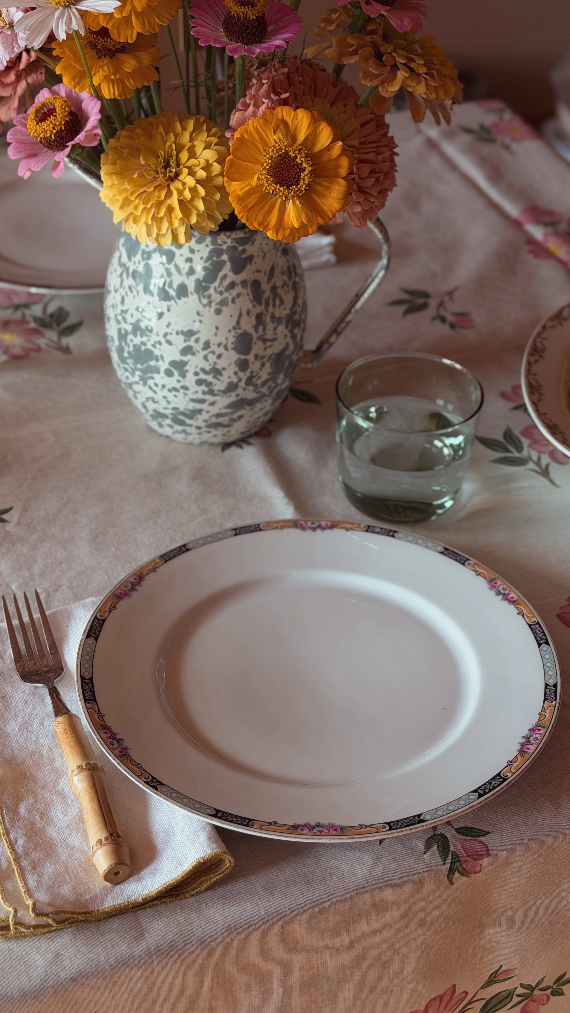 A styled tabletop setting featuring a white ceramic plate with a delicate floral gold rim, placed on a floral tablecloth next to a bamboo-handled fork and a glass of water. In the background, a speckled ceramic pitcher filled with vibrant yellow, orange, and pink flowers sits on a beige table runner.