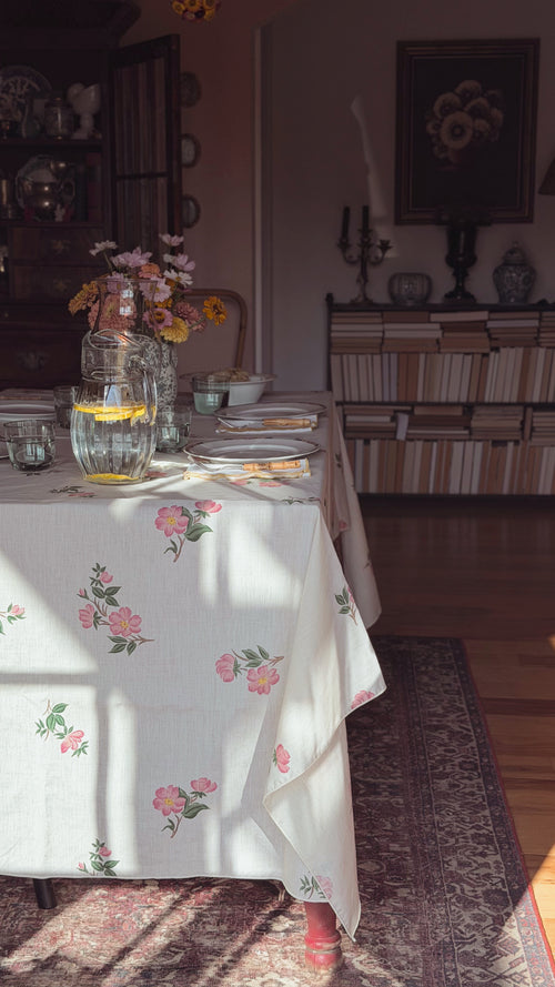 A white tablecloth with a pink and green floral print draped over a dining table in a sunlit room. The table is set with plates, a glass pitcher of lemonade, and a floral arrangement, with a patterned rug and bookshelves in the background.