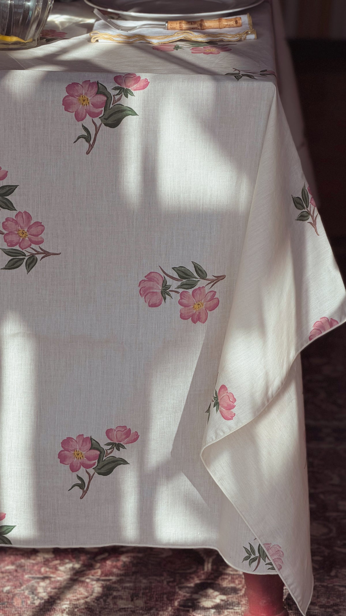 An off-white tablecloth with a delicate pink floral and green leaf pattern, draped over a table in warm, sunlit indoor light.
