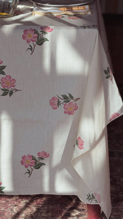 An off-white tablecloth with a delicate pink floral and green leaf pattern, draped over a table in warm, sunlit indoor light.