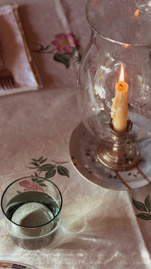 A lit cream candle in a clear glass hurricane lantern with a silver-toned base, set on a floral tablecloth next to a small glass of water.