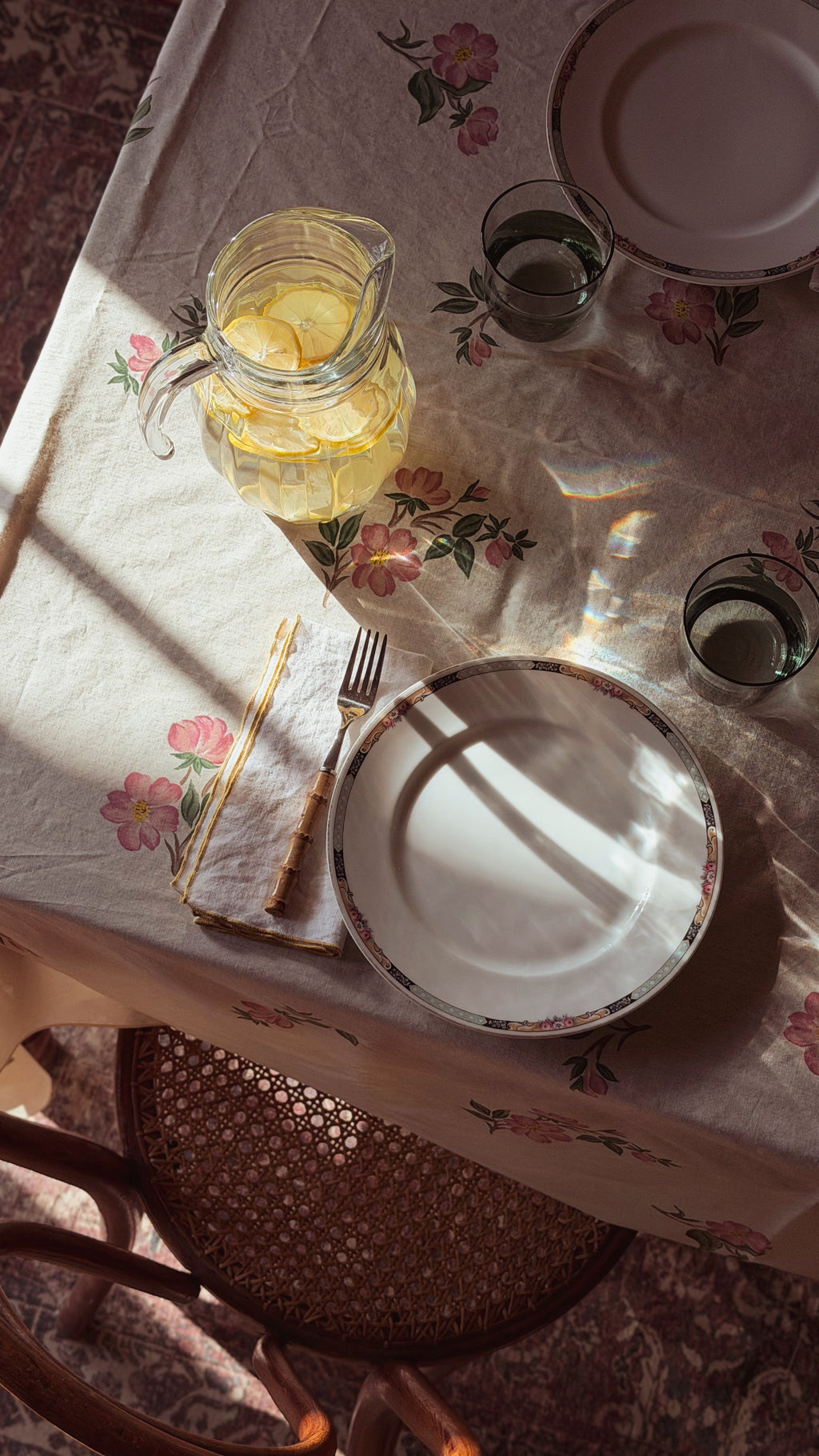 A styled dining table scene with a floral tablecloth, a glass pitcher of lemonade, plates, glasses, and cutlery arranged in warm sunlight. A woven chair is visible below the table.