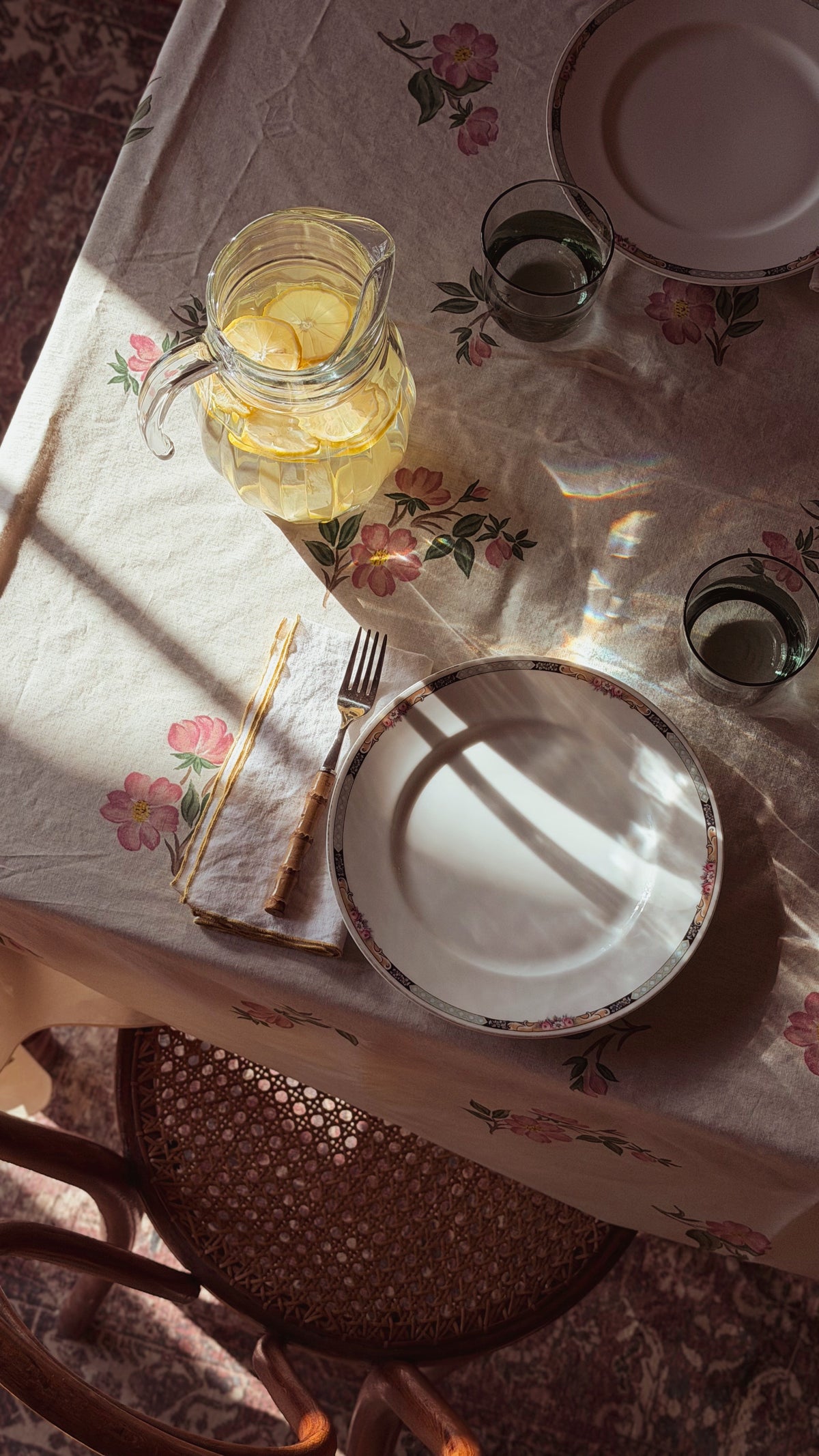 A styled dining table scene with a floral tablecloth, a glass pitcher of lemonade, plates, glasses, and cutlery arranged in warm sunlight. A woven chair is visible below the table.