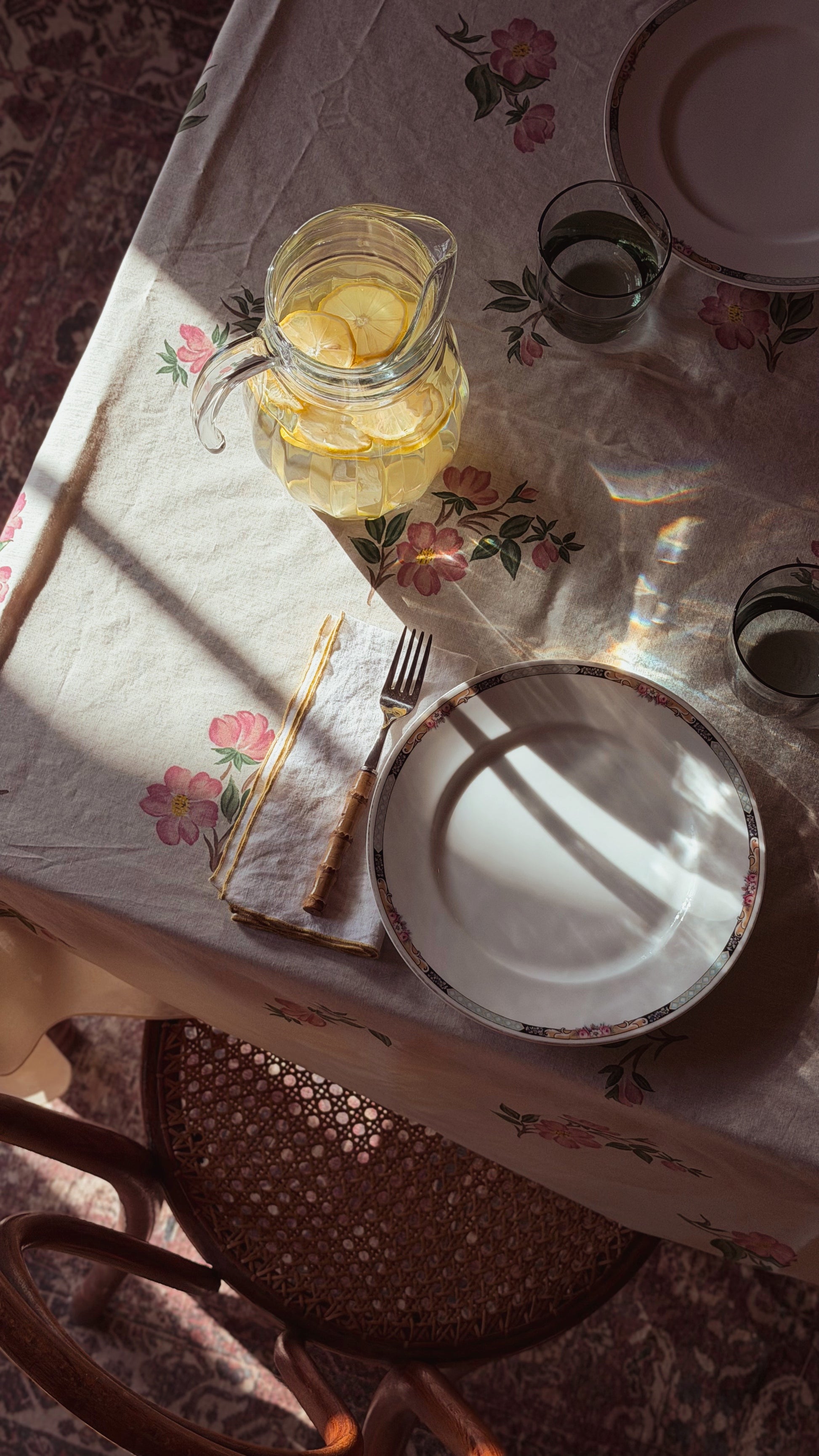 A styled tabletop scene with a floral tablecloth, a glass pitcher of lemonade with lemon slices, plates, cutlery, and two glasses, photographed in warm sunlight with strong shadows.