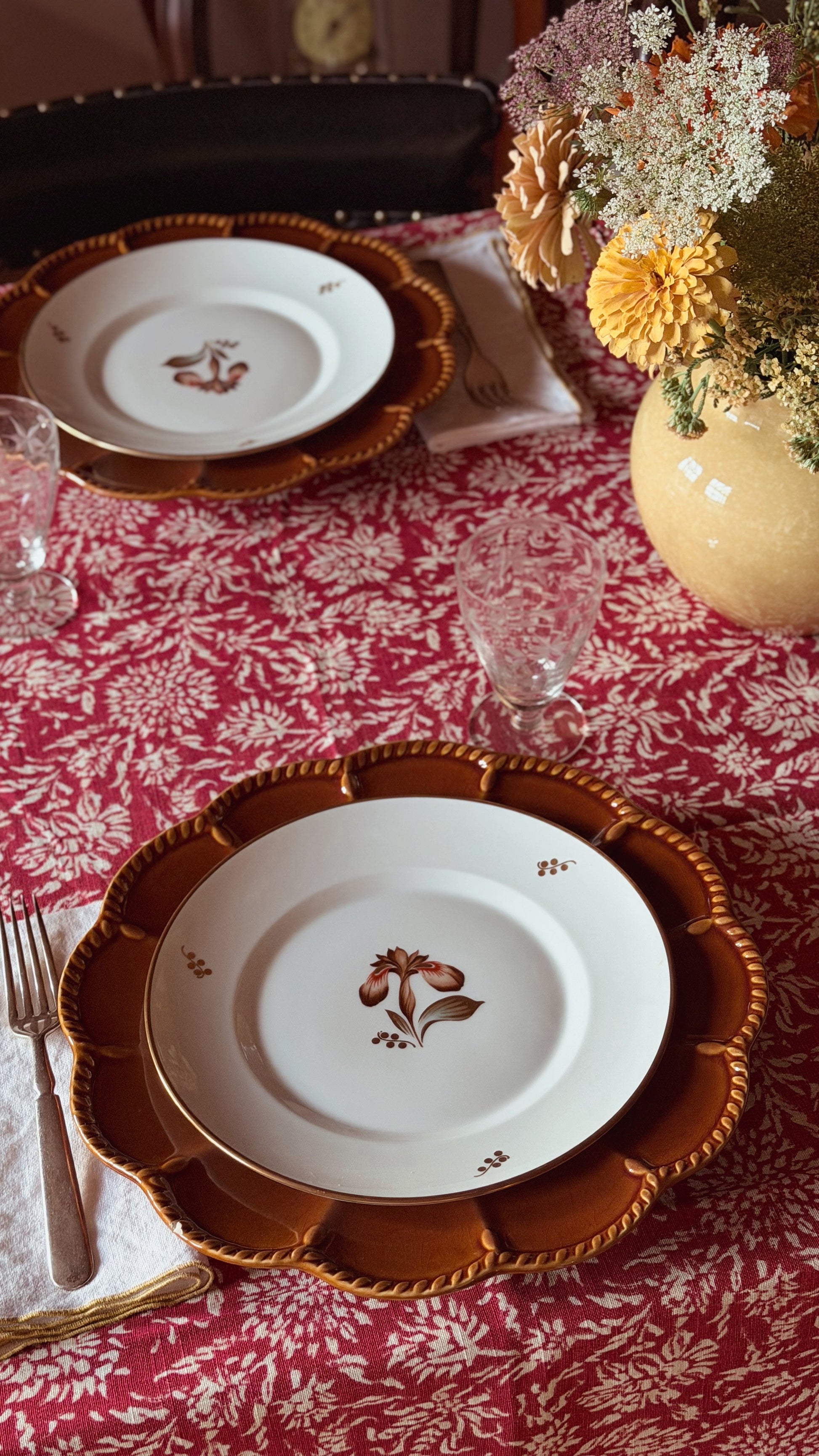 A styled table setting featuring white ceramic plates with a brown floral motif, layered over scalloped brown charger plates with gold trim, set on a red and white floral tablecloth next to a yellow vase of flowers.