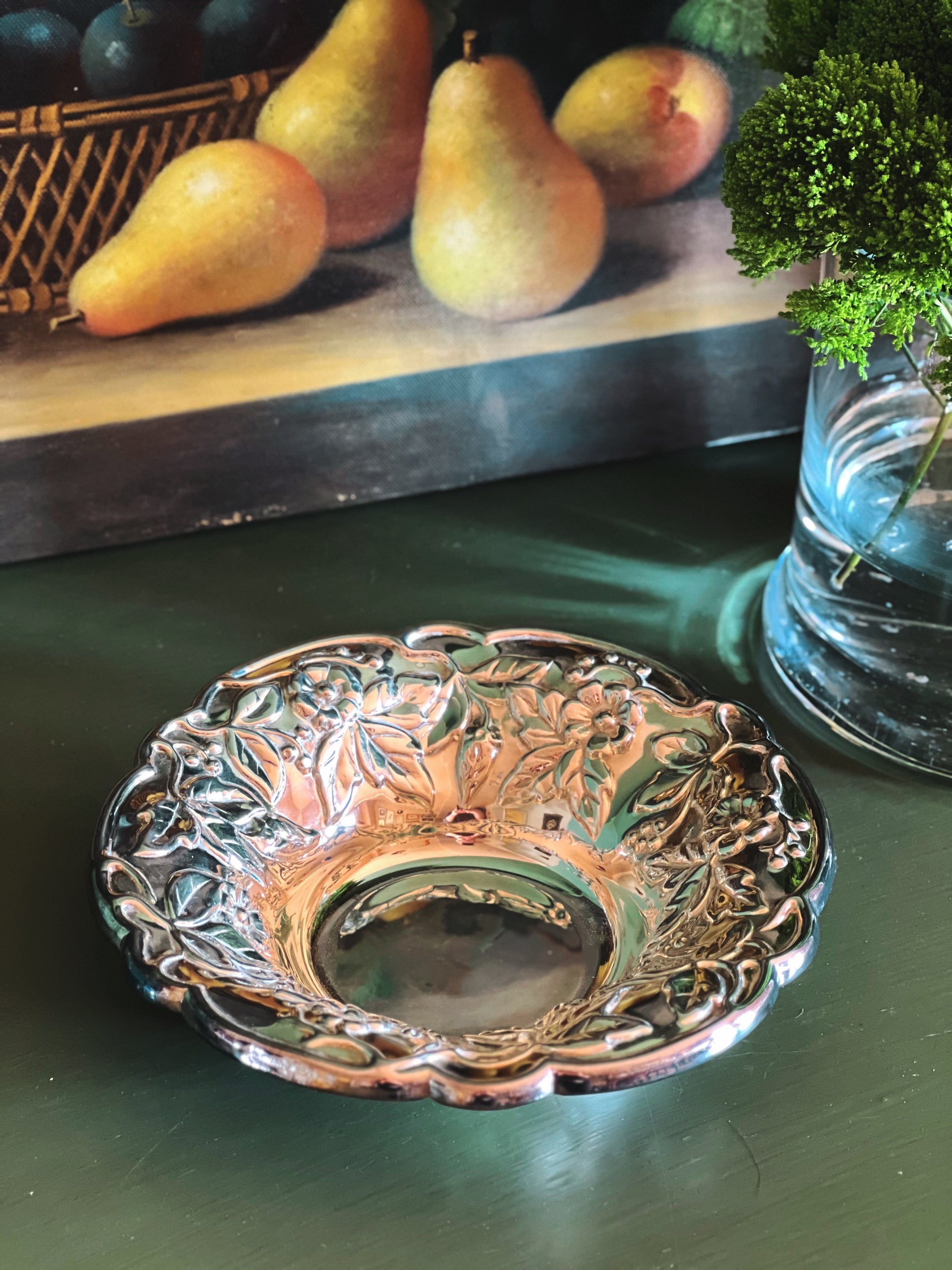 AVintage Raimond Italian Silverplate Floral Bowl, sitting on a dark green tabletop. In the background, a framed painting of pears and a glass vase with greenery are visible.