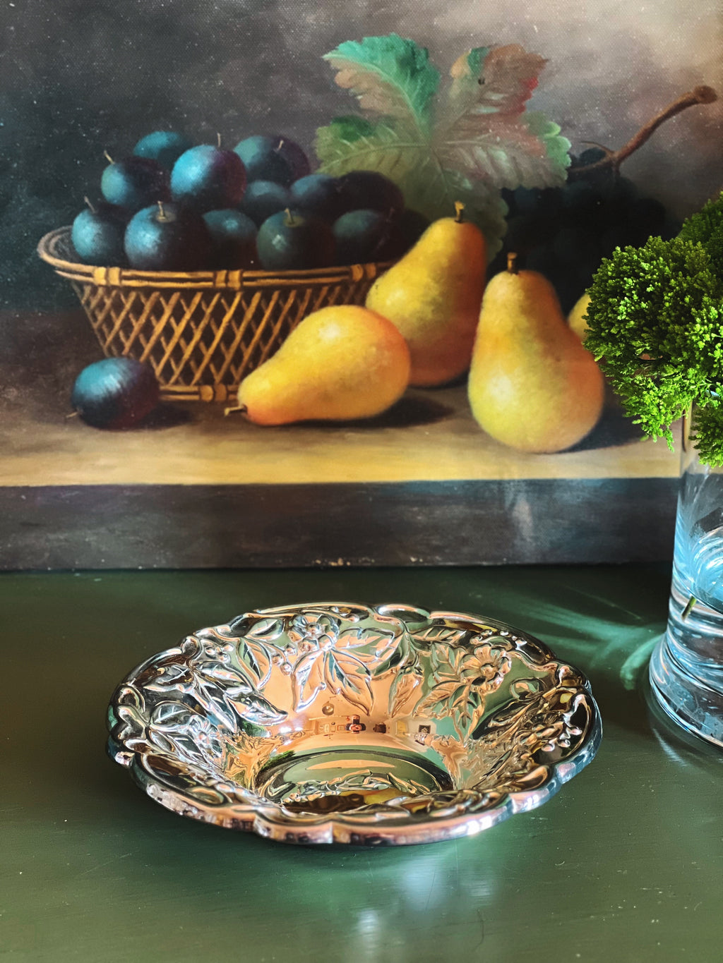 An ornate, scalloped Vintage Raimond Italian Silverplate Floral Bowl sits on a dark green table in front of a framed still life painting depicting pears and plums.