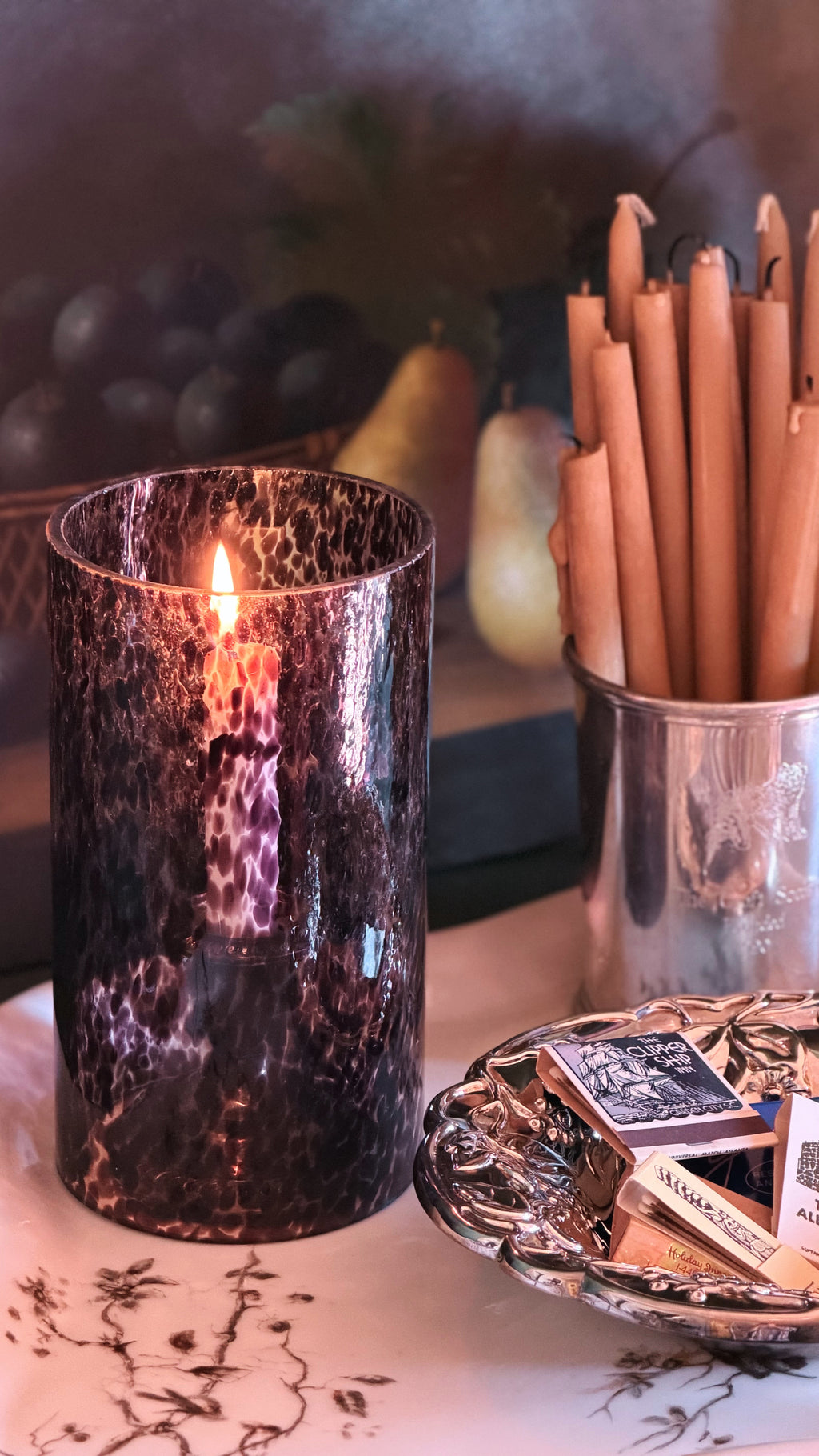 A dark, textured glass candle holder with a lit candle, styled on a table alongside a silver cup of taper candles and a small dish of matches.