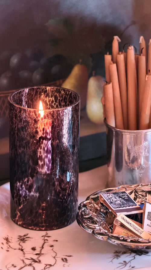 A dark, textured glass candle holder with a lit candle, styled on a table alongside a silver cup of taper candles and a small dish of matches.
