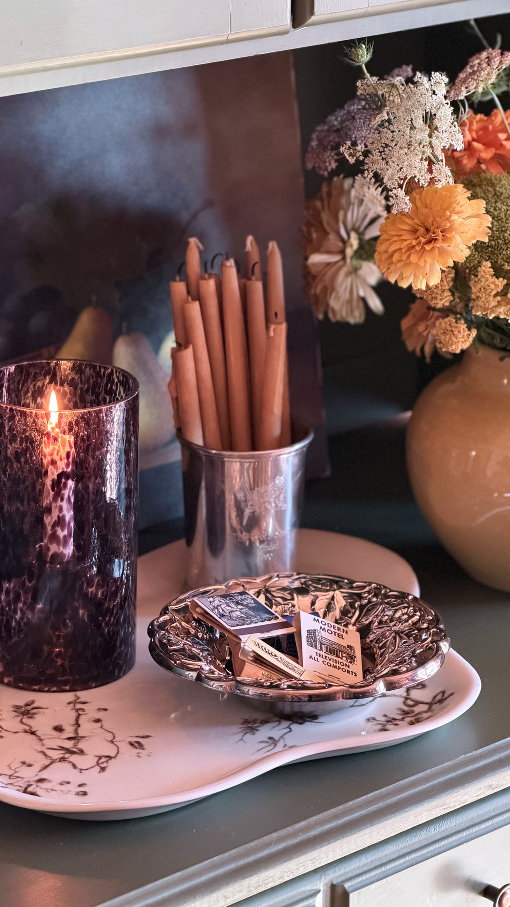A styled tabletop scene with a lit dark amber candle in a textured holder, a metal cup holding wooden wicks, a small decorative dish with matchbooks, and a floral arrangement in the background.