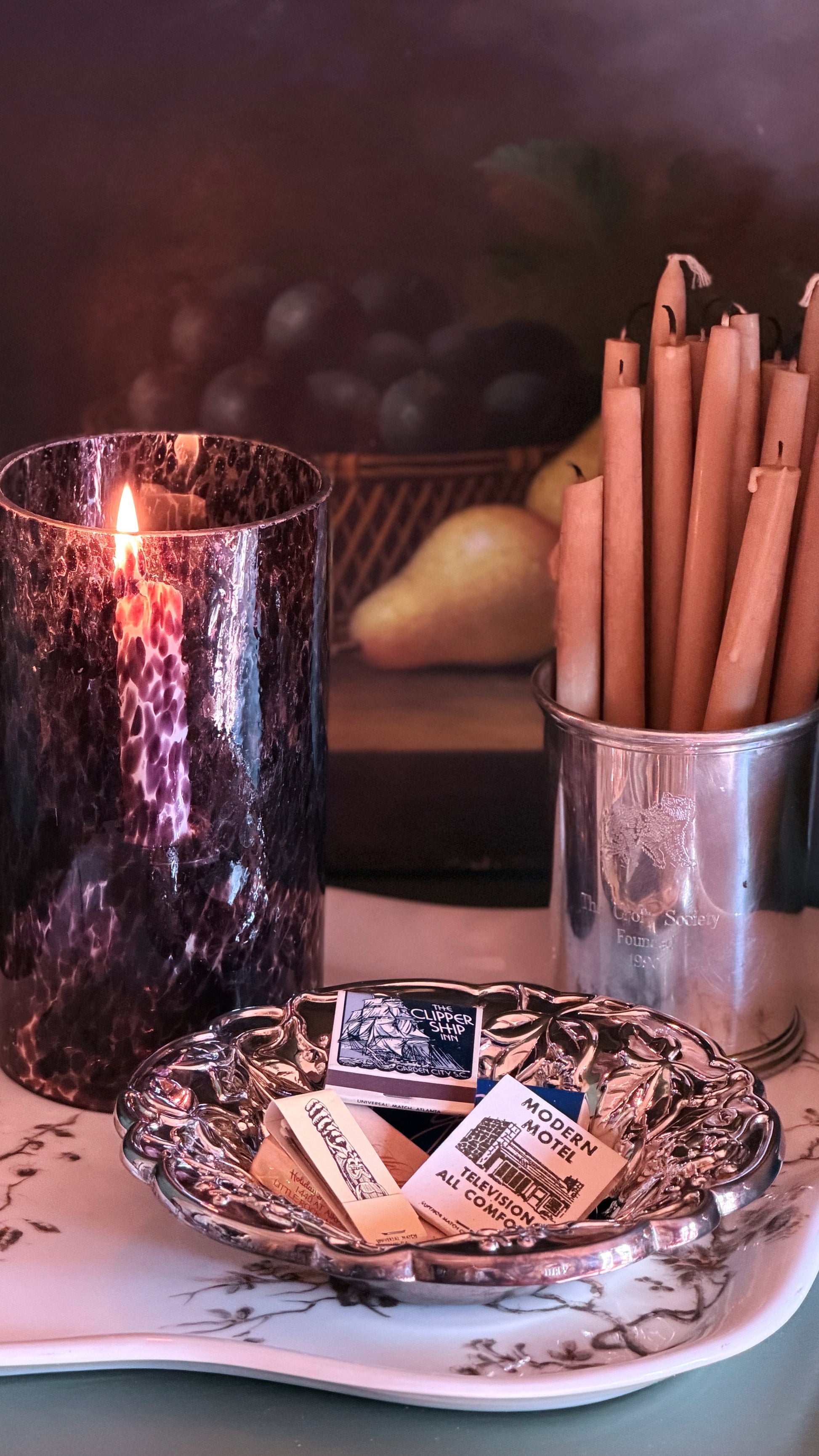A styled tabletop scene with a lit dark purple marbled candle in a tall glass holder, a metal cup filled with taper candles, a small silver dish holding matchbooks, and a pear in the background.
