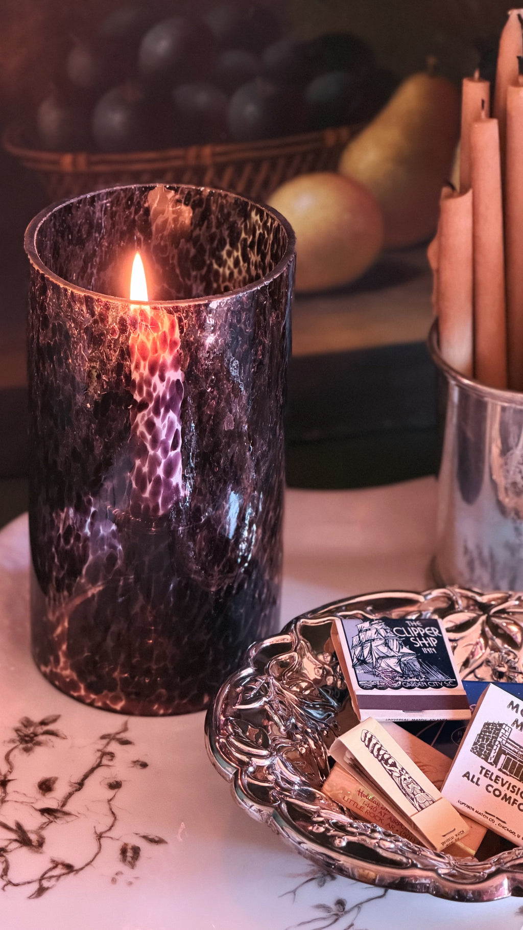 A lit candle in a dark, textured cylindrical holder with a metallic bronze and black marbled pattern, styled on a table next to a silver dish with matchboxes.