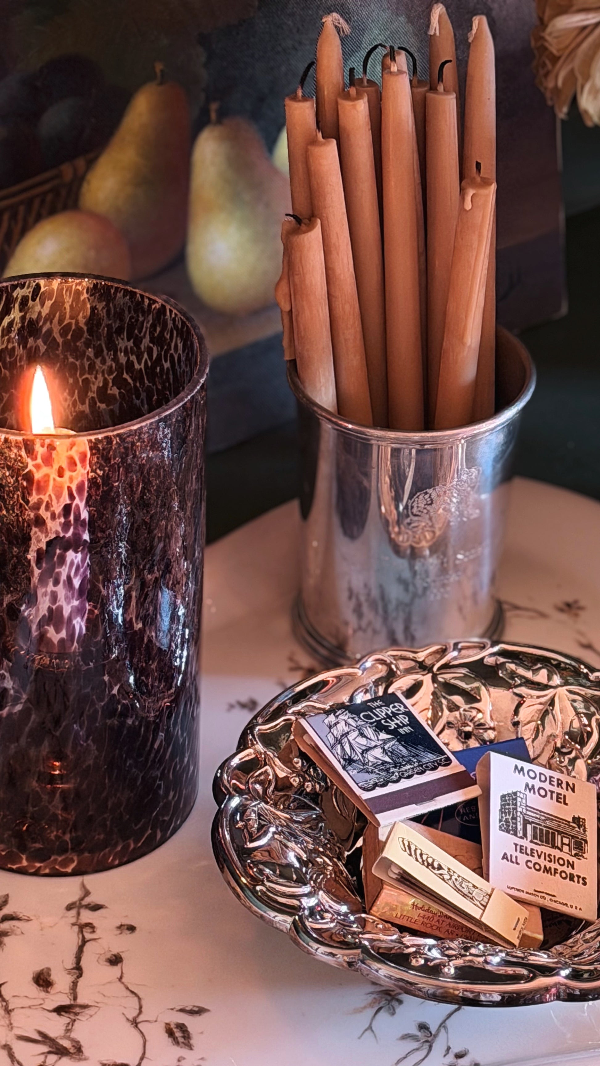 A styled tabletop scene with a lit candle in a dark hammered glass holder, a metal cup holding several wooden taper candles, and a decorative silver tray holding matchboxes and a small card reading 'Modern Comfort'.