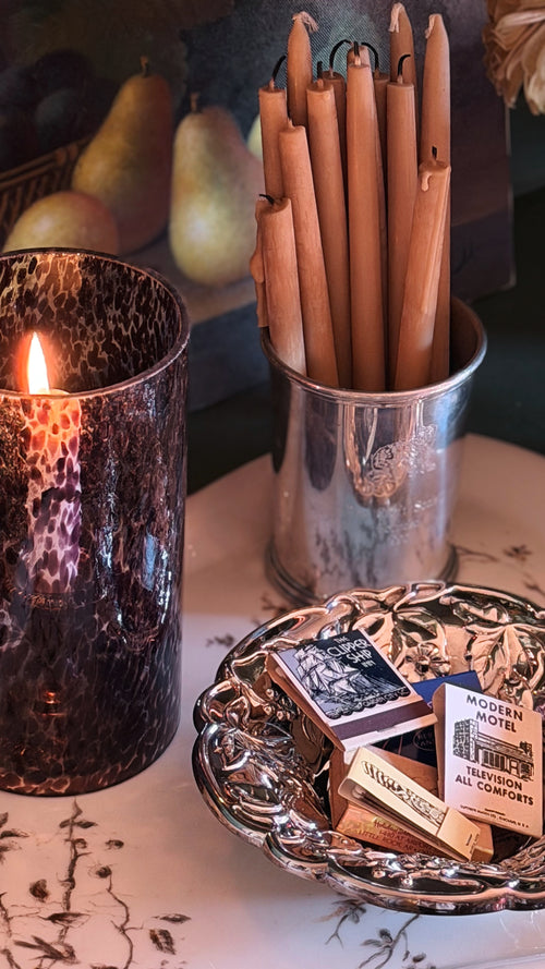 A styled tabletop scene with a lit candle in a dark hammered glass holder, a metal cup holding several wooden taper candles, and a decorative silver tray holding matchboxes and a small card reading 'Modern Comfort'.
