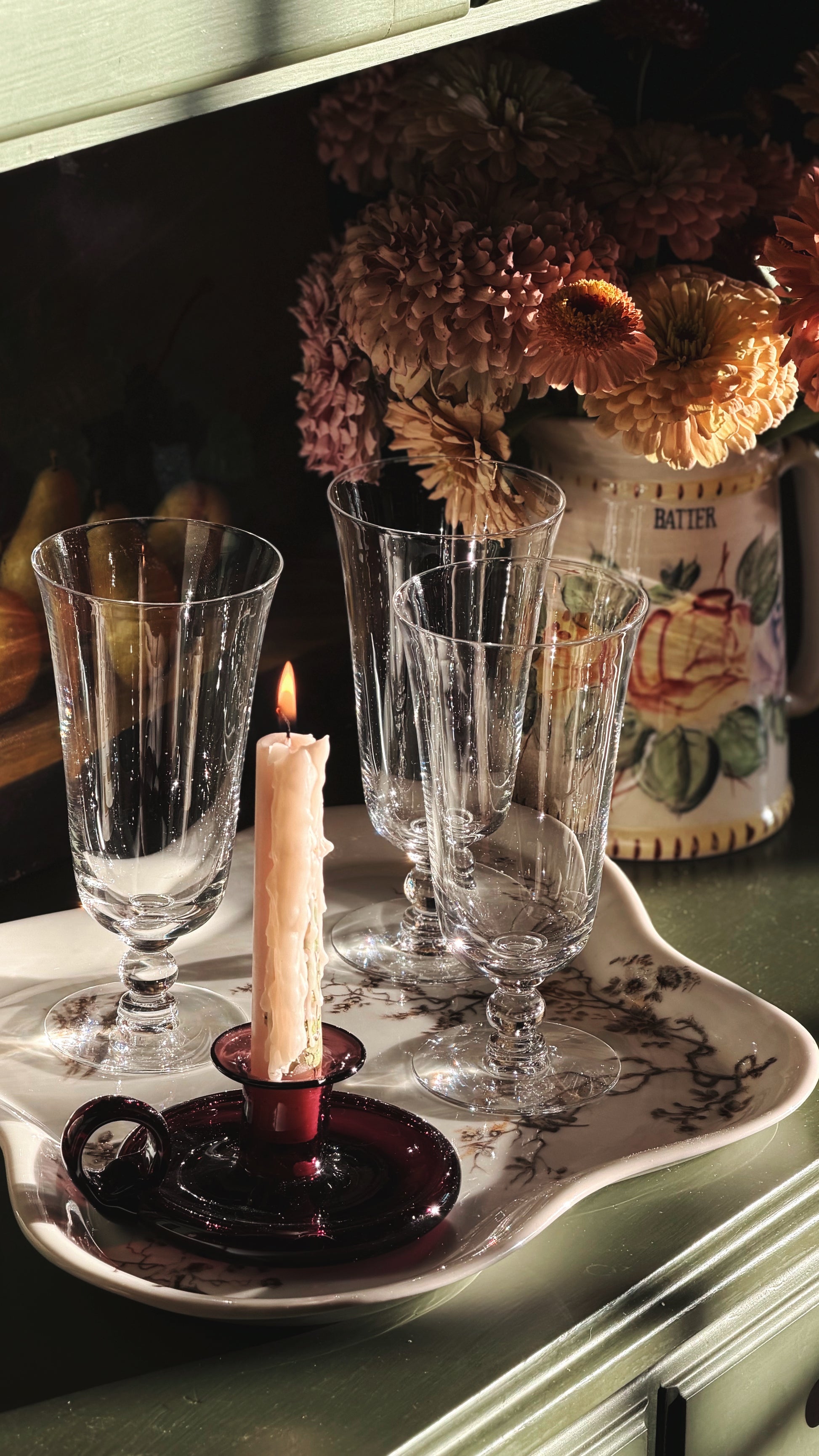 A set of four clear ribbed glass goblets arranged on a white ceramic tray with a dark purple candle holder and lit candle, with dried flowers in the background.