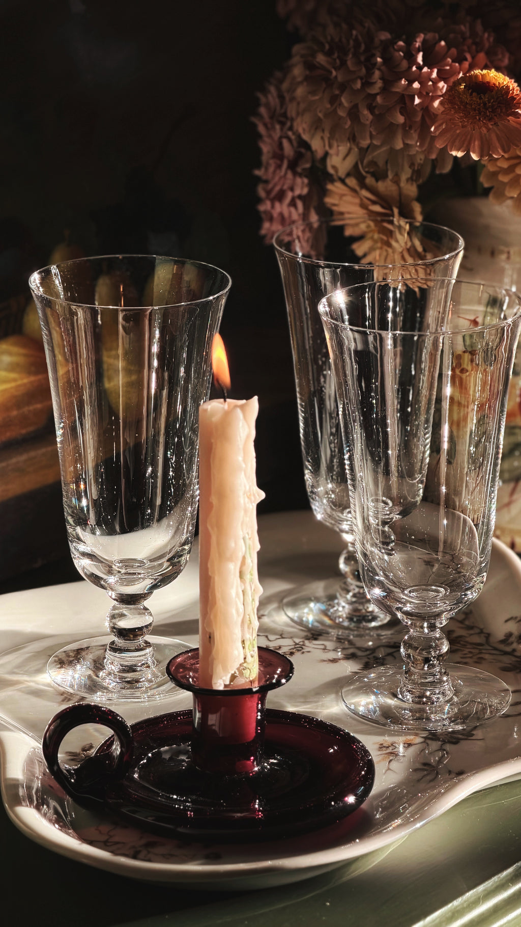 A set of clear ribbed glass goblets and a dark purple glass candlestick holder with a lit white candle, arranged on a white tray with dried flowers in the background.