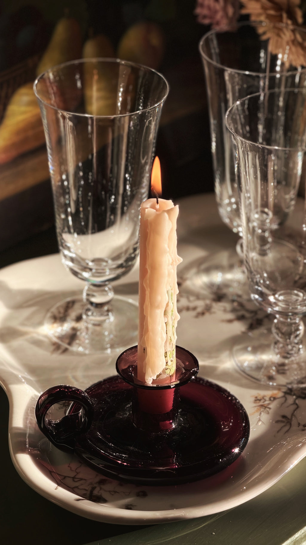 A lit cream-colored candle in a dark purple glass holder, set on a white ceramic tray with floral detailing, surrounded by several clear glass goblets in a dimly lit setting.