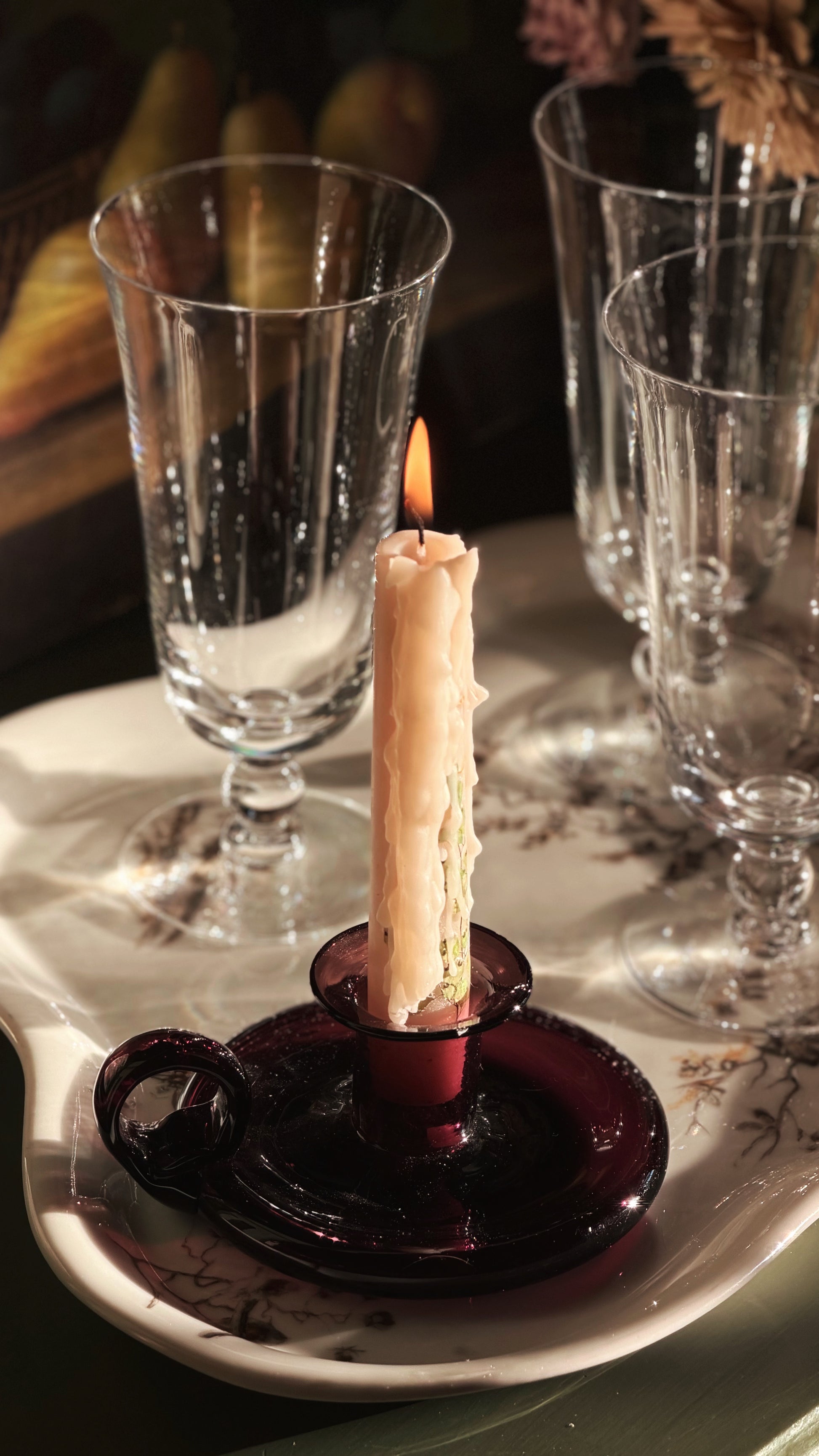 A lit cream-colored candle in a dark purple glass holder, set on a white ceramic tray with floral detailing, surrounded by several clear glass goblets in a dimly lit setting.