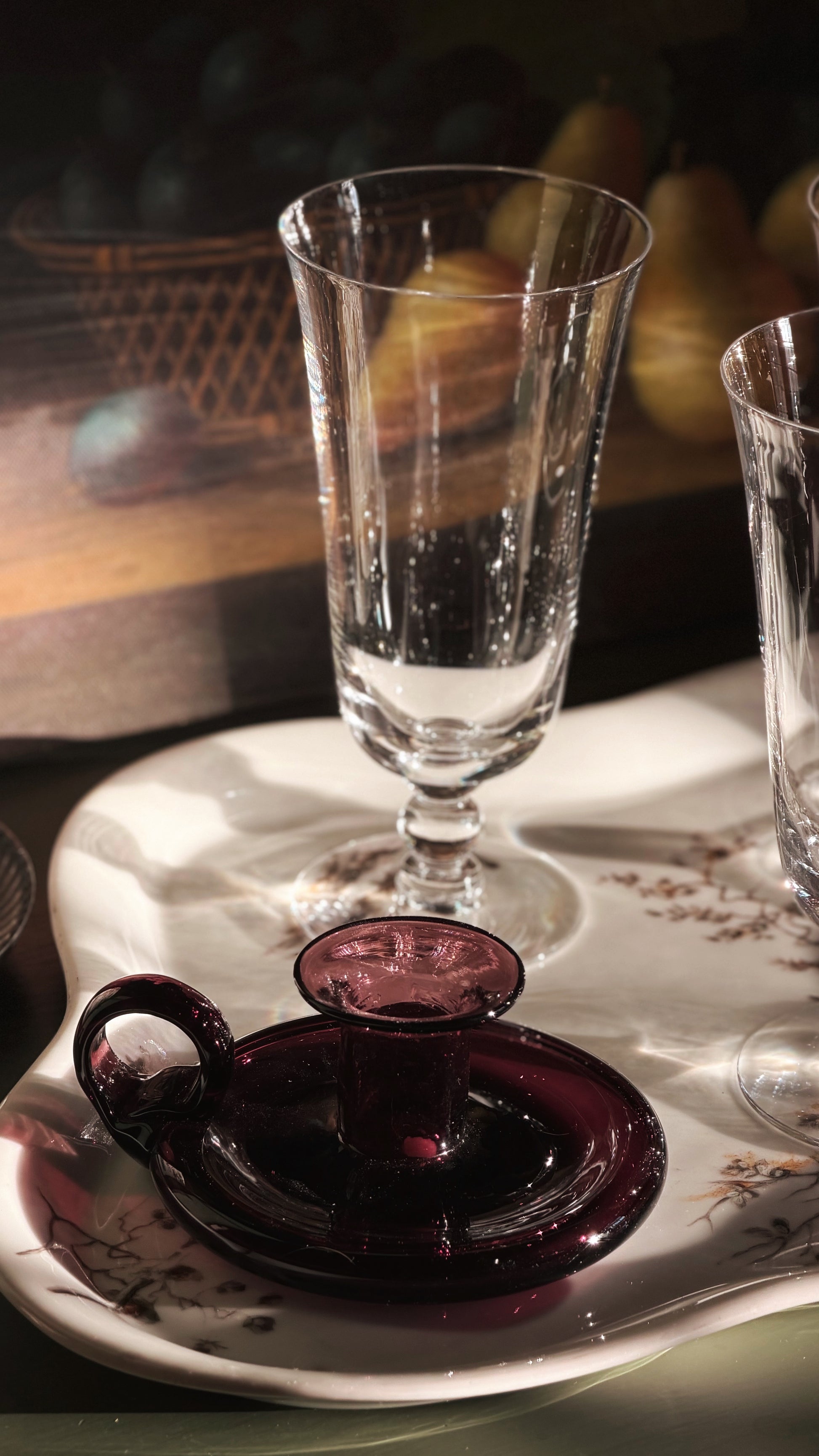 A clear glass goblet and a dark purple glass candle holder set on a white floral ceramic tray, with warm, moody lighting in a dimly lit interior setting.