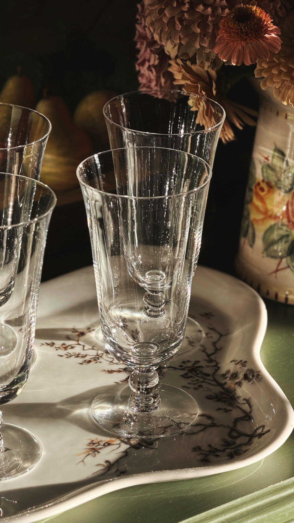Clear glass crystal stemware with flared rims and ribbed sides, arranged on a decorative white tray with floral detailing, in warm dim lighting with dried flowers in the background.