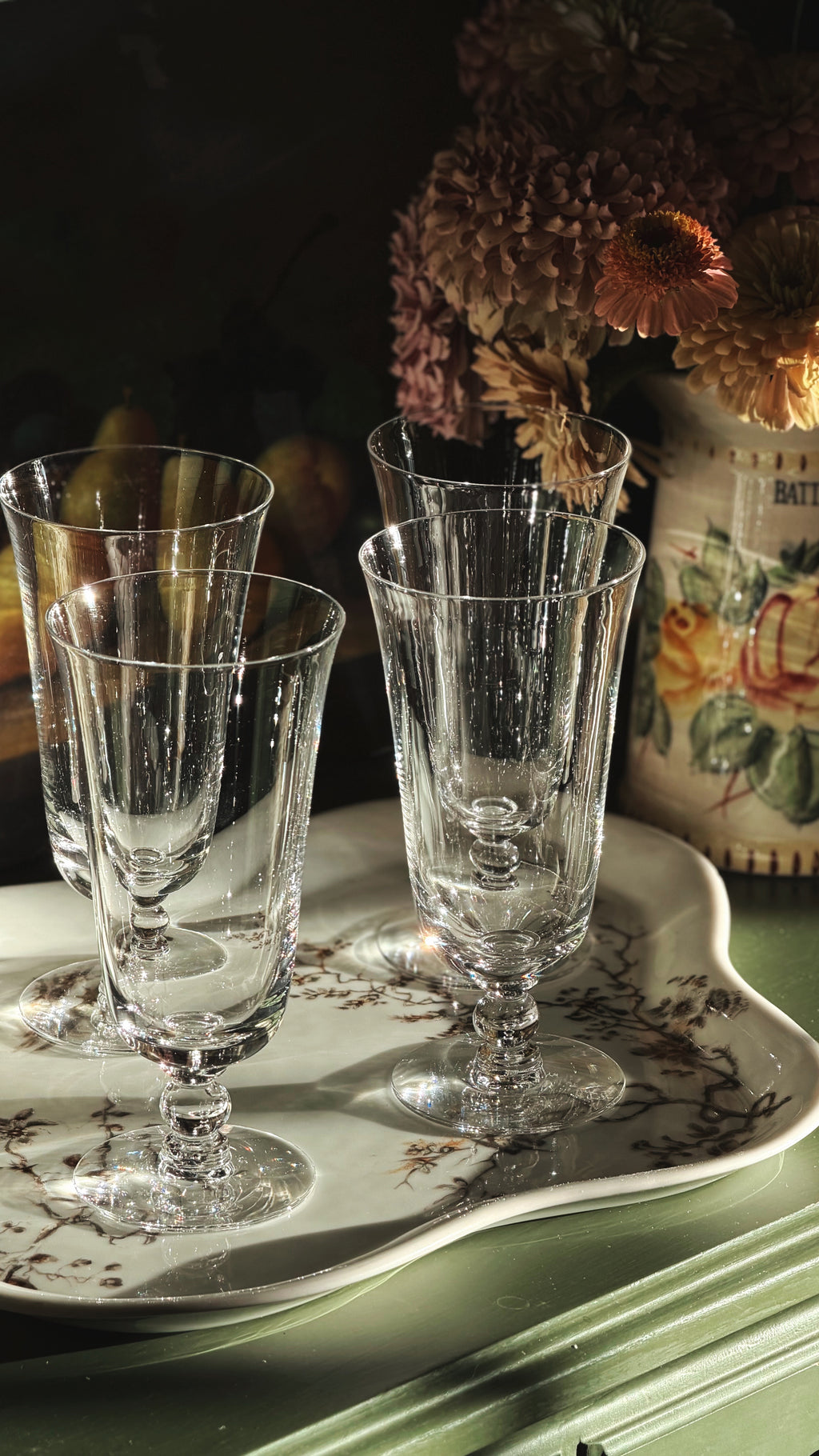 Four clear crystal iced tea glasses arranged on a white ceramic tray with a floral border, set on a green wooden surface with a vase of dried flowers in the background.