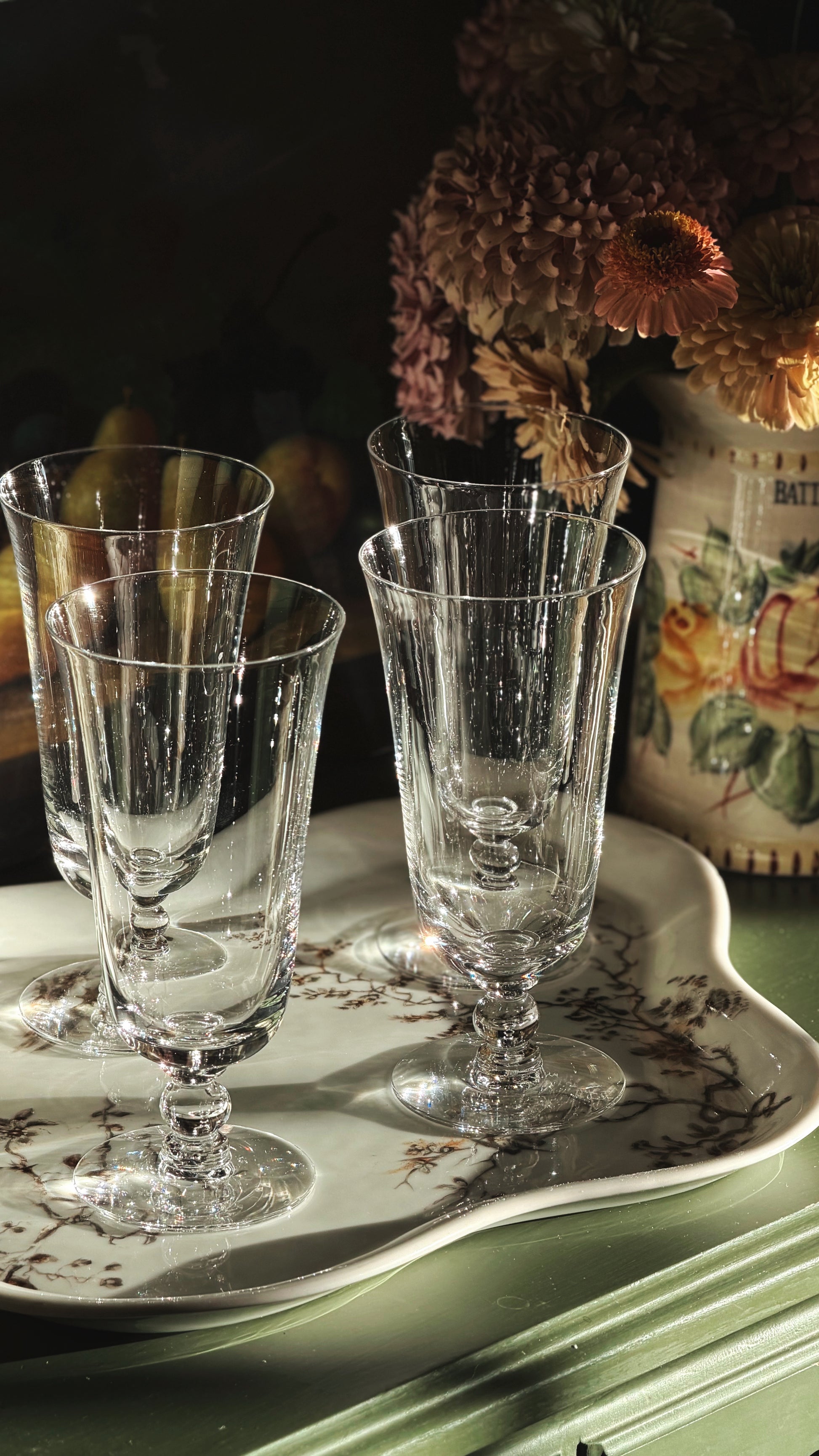 Four clear crystal iced tea glasses arranged on a white ceramic tray with a floral border, set on a green wooden surface with a vase of dried flowers in the background.