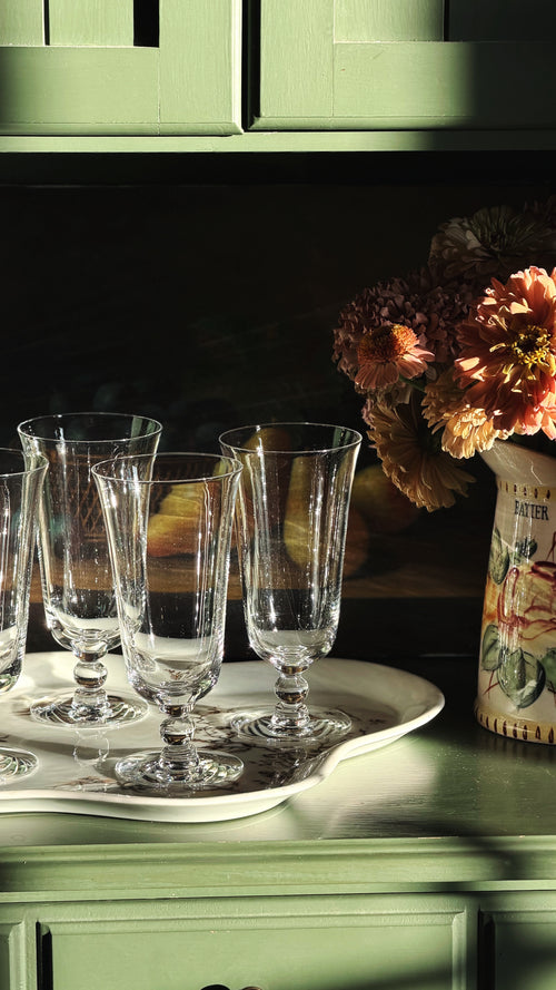 A set of clear glass tulip-shaped wine glasses arranged on a white oval tray on a green wooden cabinet, with warm sunlight casting shadows and a vase of flowers in the background.