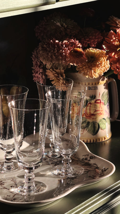 A set of clear glass stemware glasses arranged on a decorative white tray with floral detailing, set against a dark background with dried flowers and a ceramic pitcher.