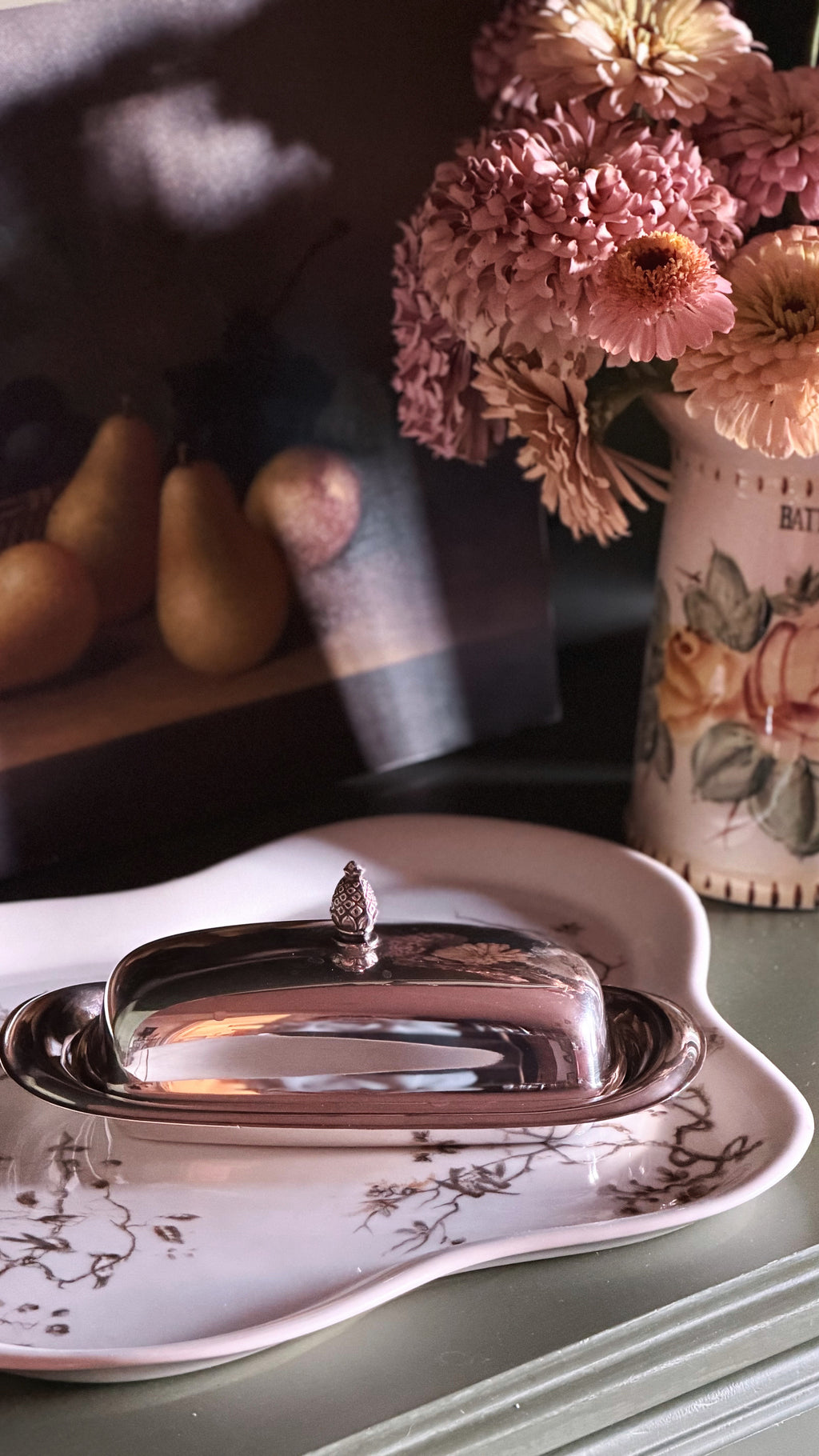 A polished silver butter dish with a decorative finial lid, resting on a white ceramic plate with a floral pattern, set against a dark background with dried flowers and pears.