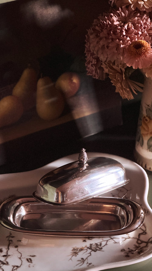 A polished silver metal butter dish with its lid propped open on a white ceramic plate with delicate brown floral detailing, set against a dark, moody background with soft-focus flowers and fruit.