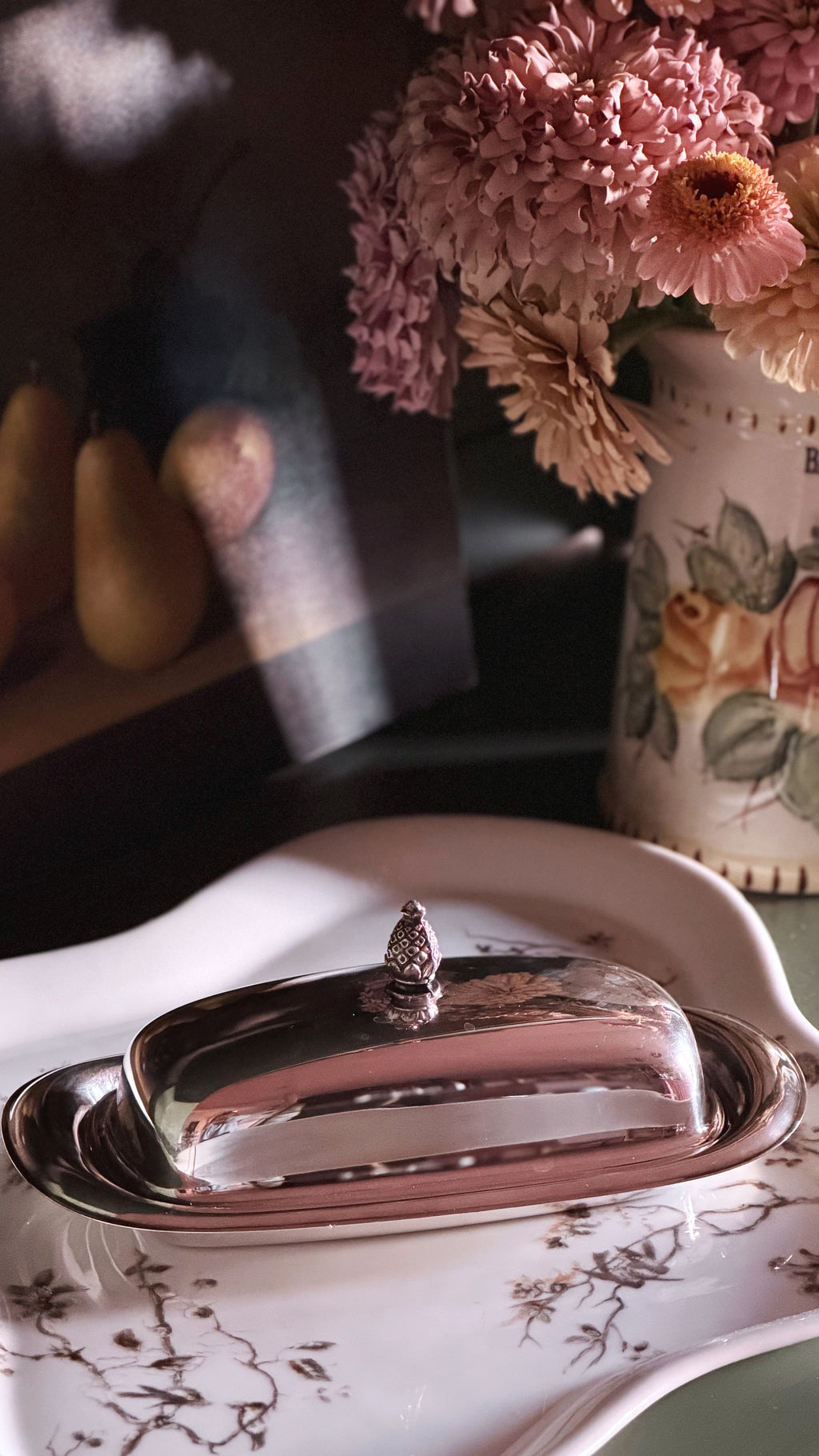 A polished silver butter dish with a domed lid and small finial, set on a white floral ceramic platter. The scene is styled with pink flowers in a patterned vase and soft warm shadows.