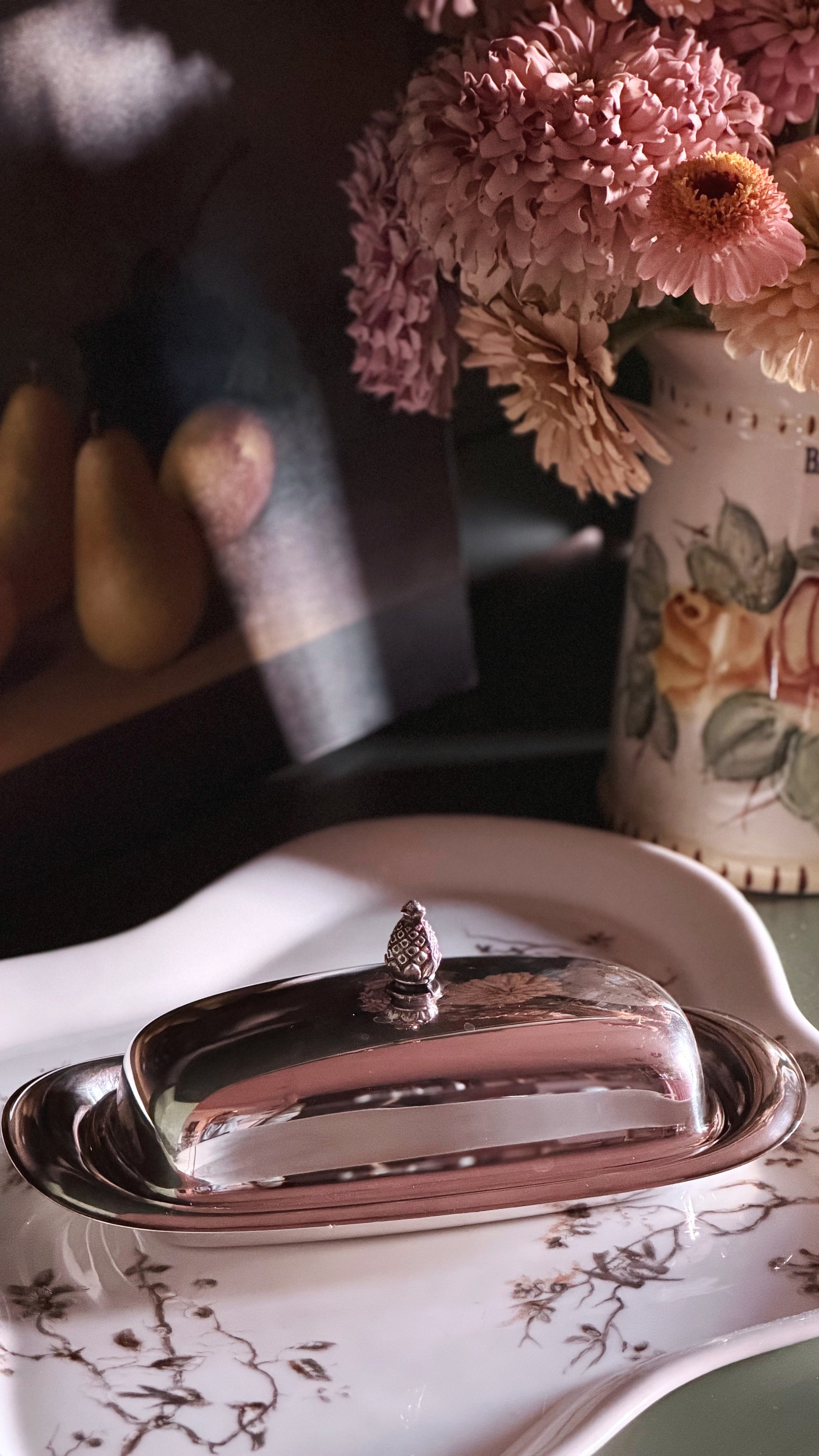 A polished silver butter dish with a domed lid and small finial, set on a white floral ceramic platter. The scene is styled with pink flowers in a patterned vase and soft warm shadows.