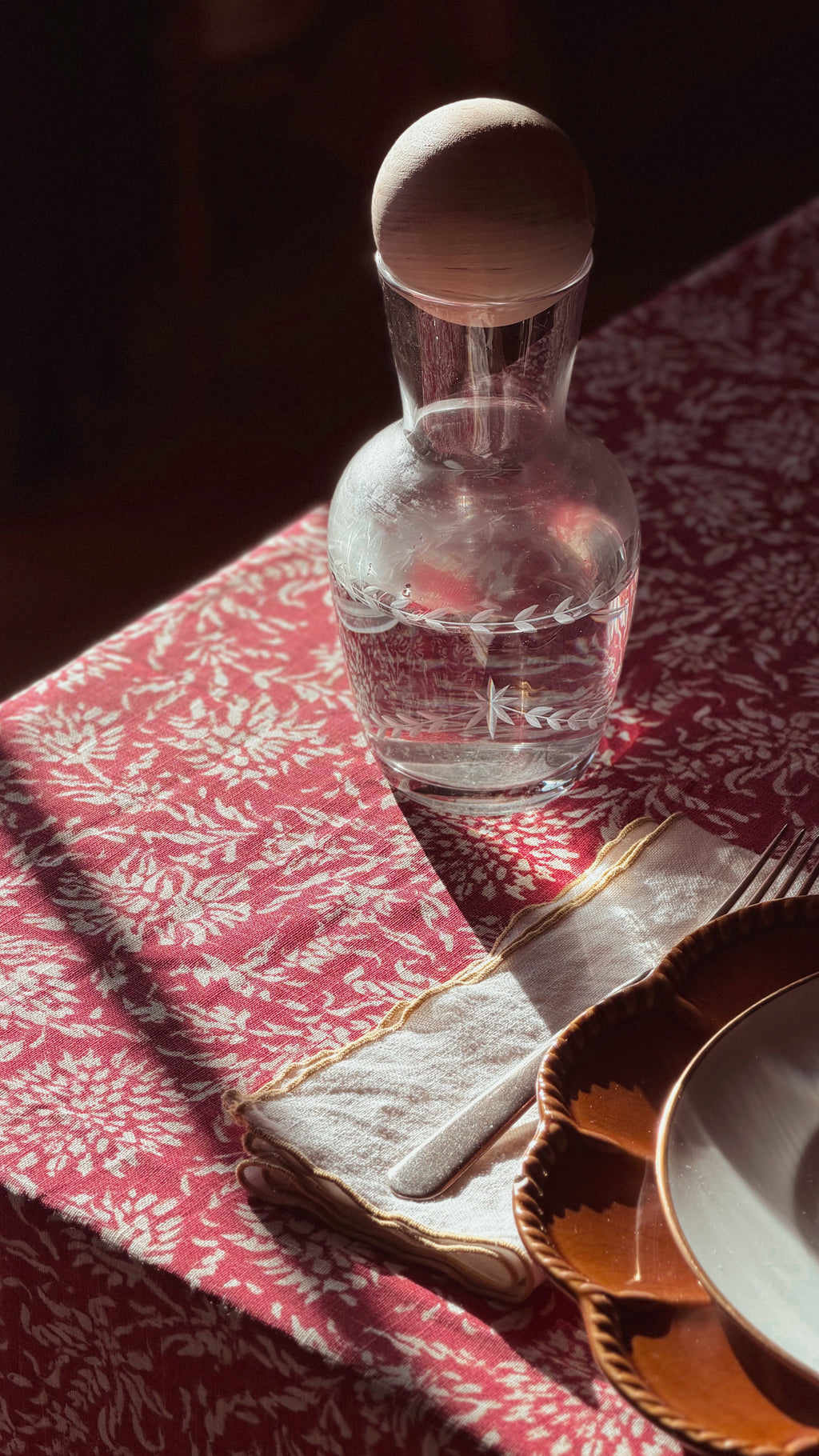 A clear glass decanter with a wooden stopper sits on a red and white patterned tablecloth next to a folded cream napkin, gold charger plate, and white plate in warm sunlight.