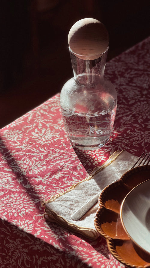 A clear glass decanter with a wooden stopper sits on a red and white patterned tablecloth next to a folded cream napkin, gold charger plate, and white plate in warm sunlight.