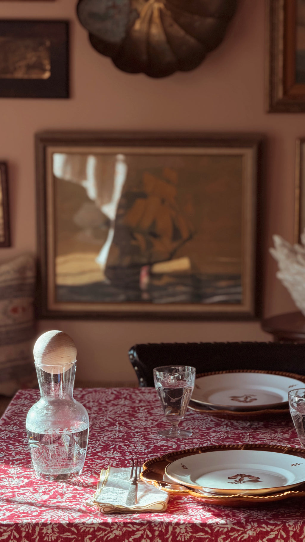 A styled dining table in a warm, rustic room setting, featuring a red and white patterned tablecloth, white plates with gold trim, a clear glass carafe, and a glass water glass. A framed painting hangs on the dusty rose wall in the background.