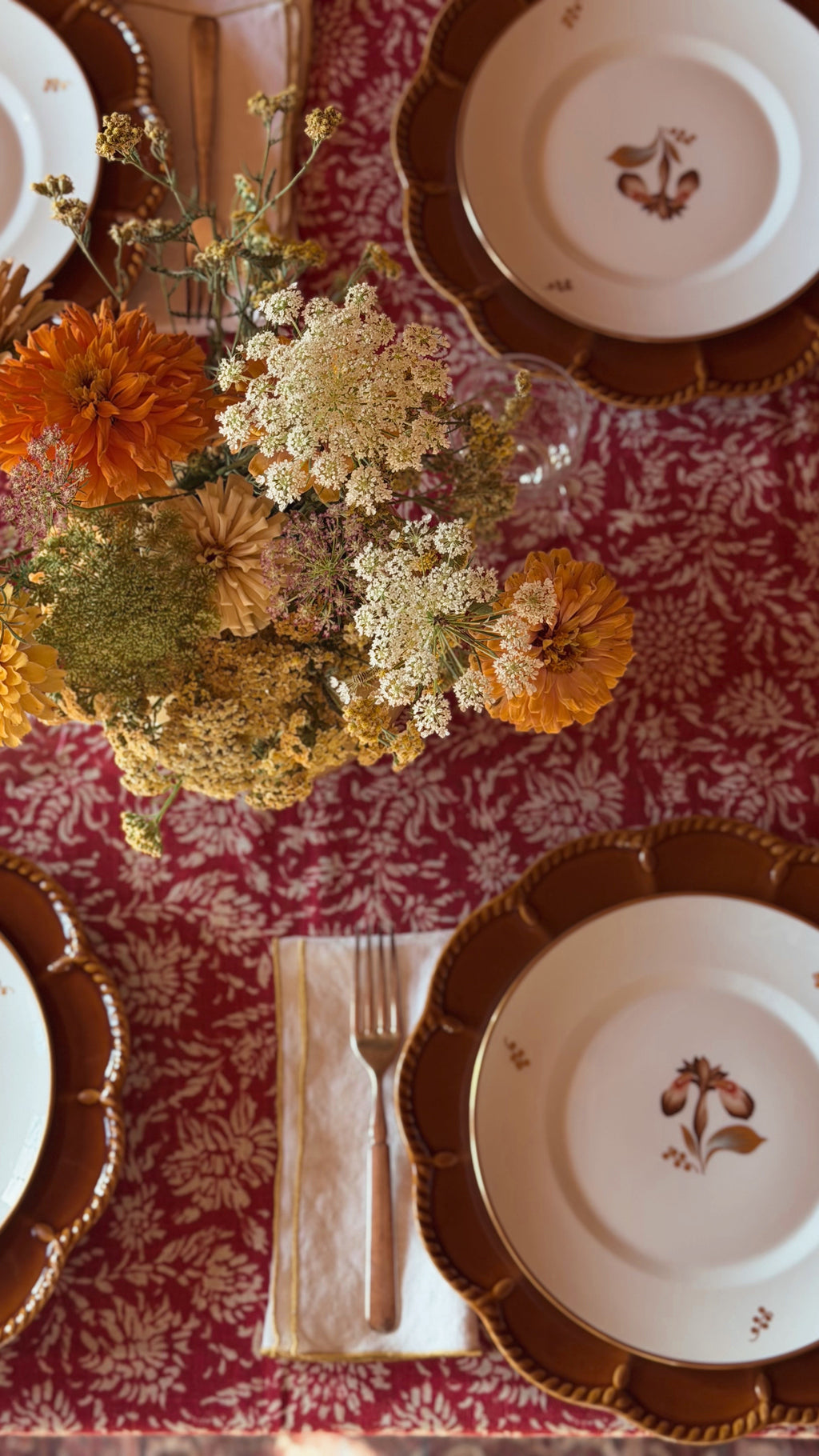 An overhead view of a styled dining table setting featuring a red and gold damask tablecloth, white ceramic plates with gold leaf scalloped edges and botanical motifs, a gold fork on a cream napkin, and a central dried flower arrangement.