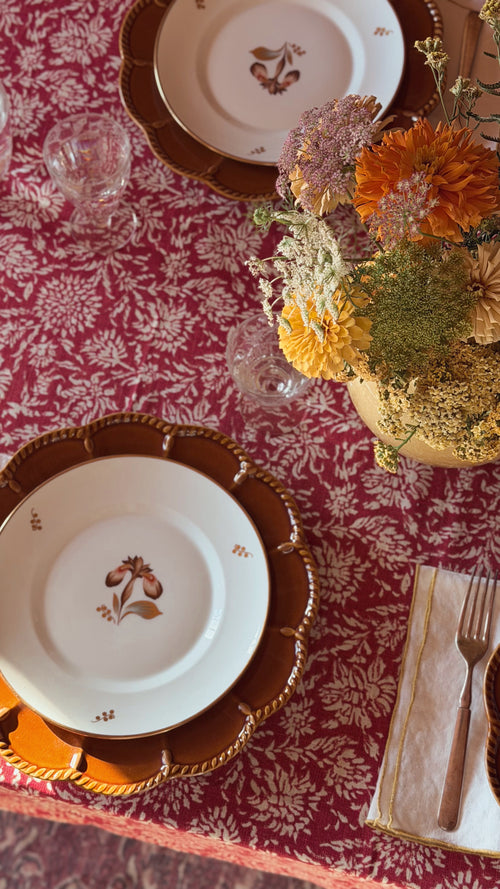 A styled table setting featuring white porcelain plates with brown floral motifs and scalloped brown chargers, set on a red damask tablecloth next to a gold-rimmed napkin with a fork and a floral centerpiece.