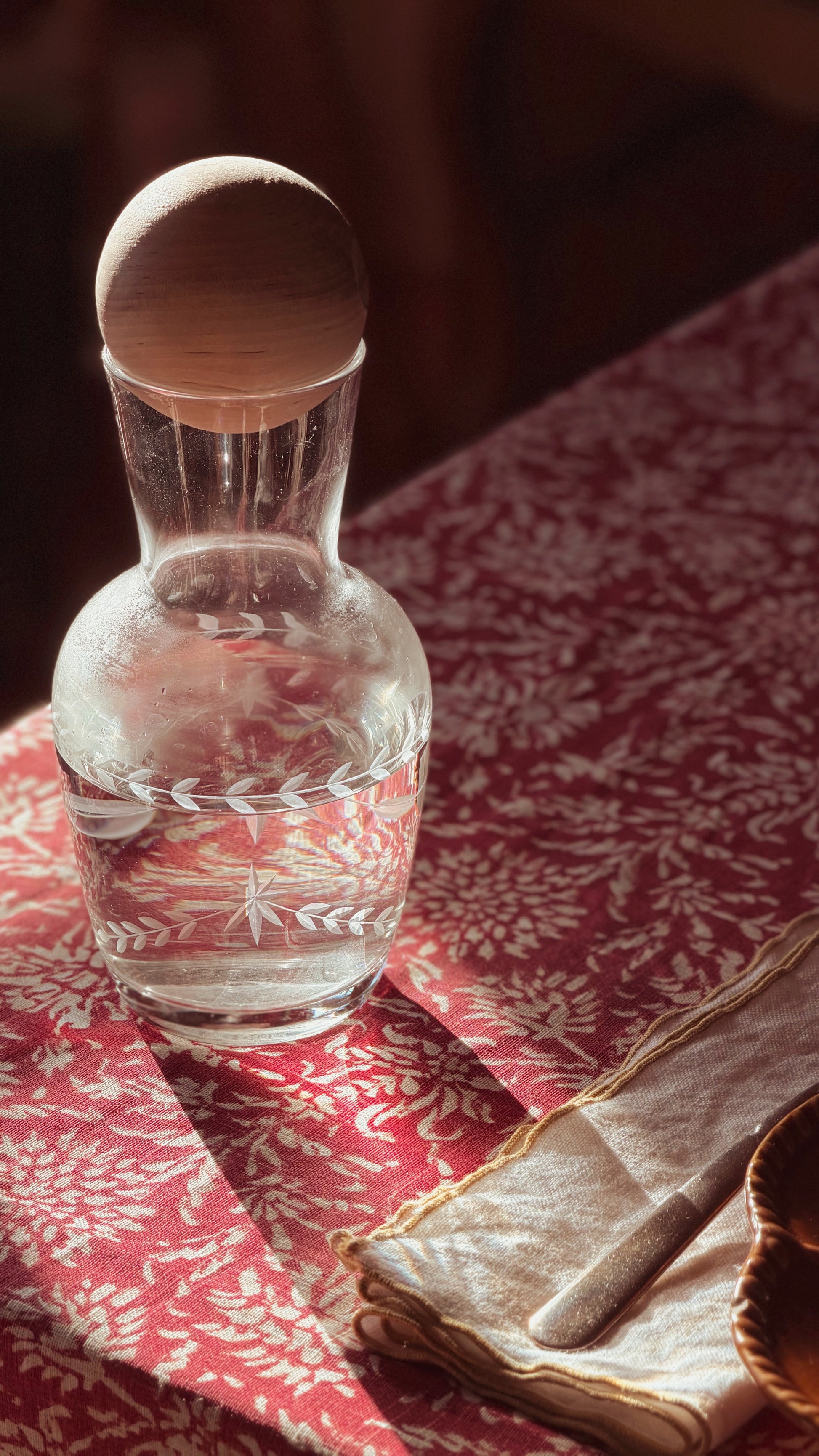 A clear glass decanter with a round wooden stopper, filled with water and decorated with a white leaf pattern near the base, resting on a red and white patterned tablecloth next to a folded napkin.