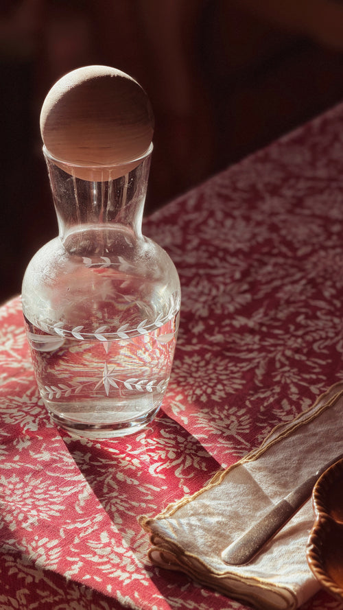 A clear glass decanter with a round wooden stopper, filled with water and decorated with a white leaf pattern near the base, resting on a red and white patterned tablecloth next to a folded napkin.