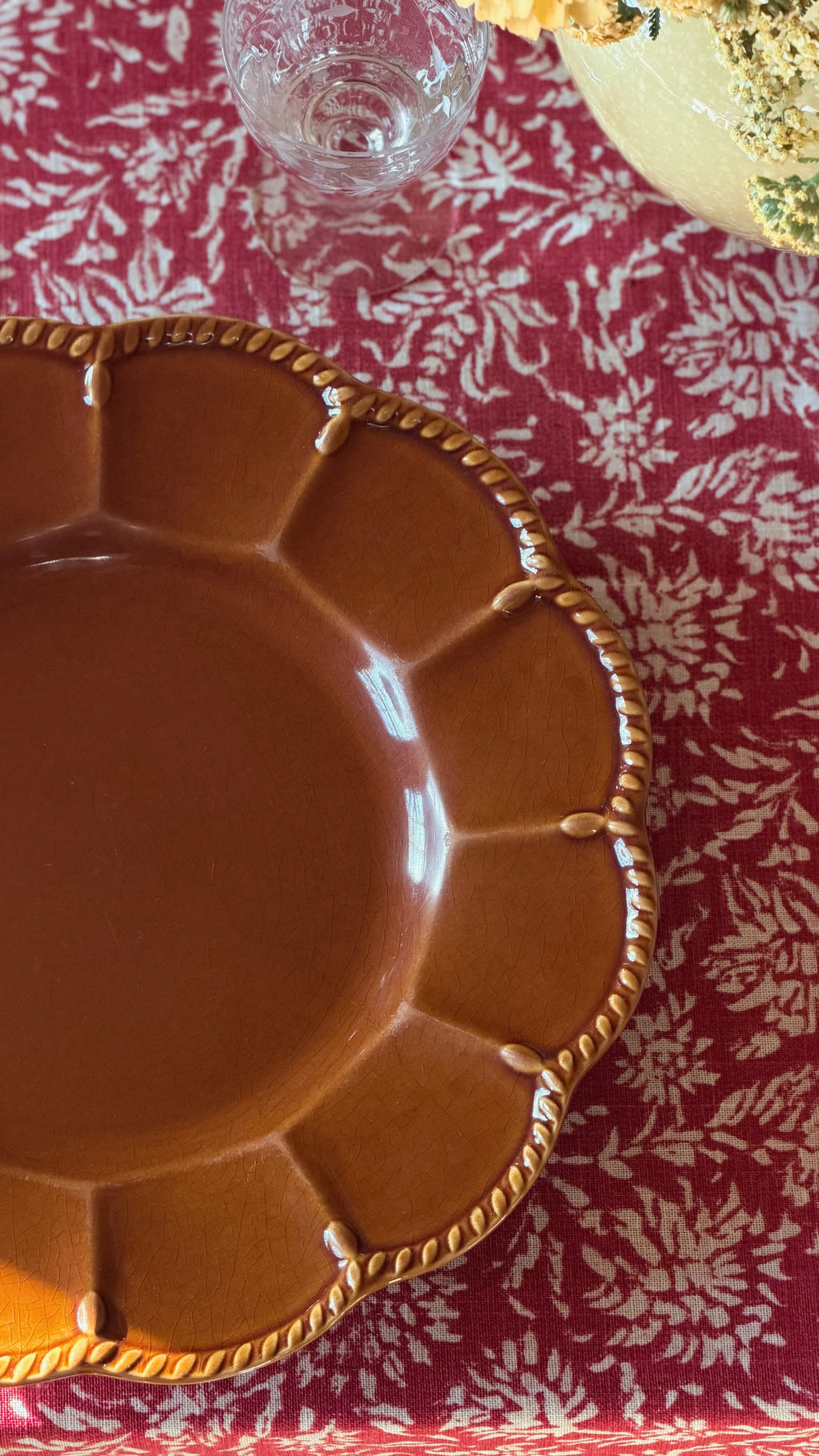 A Vintage Certified International Florentine Brown Dinner Plate by Kari Walmsley with a scalloped rim and decorative rope-like edge detailing, shown on a red floral tablecloth.