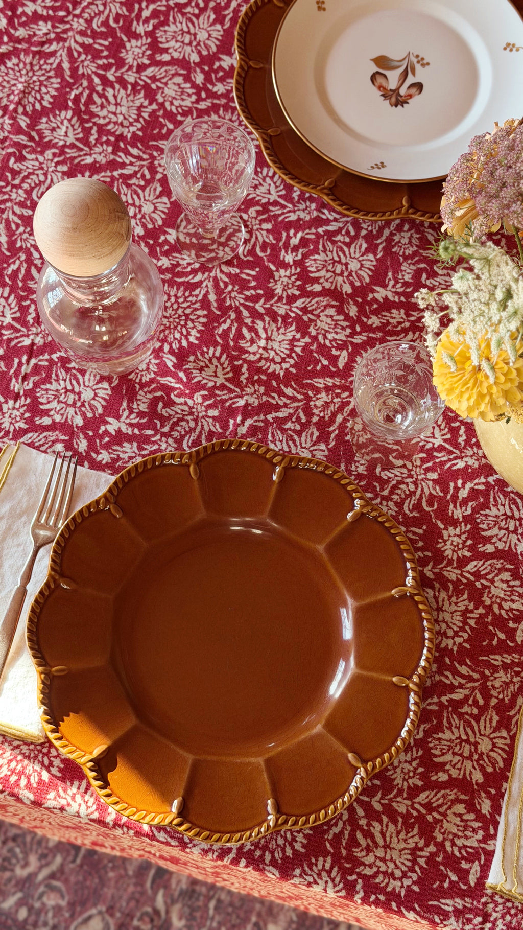 An overhead view of a styled table setting featuring aVintage Certified International Florentine Brown Dinner Plate by Kari Walmsley with a scalloped rim and gold rope detailing, placed on a red and white floral tablecloth alongside glassware, a small bottle, and floral accents.